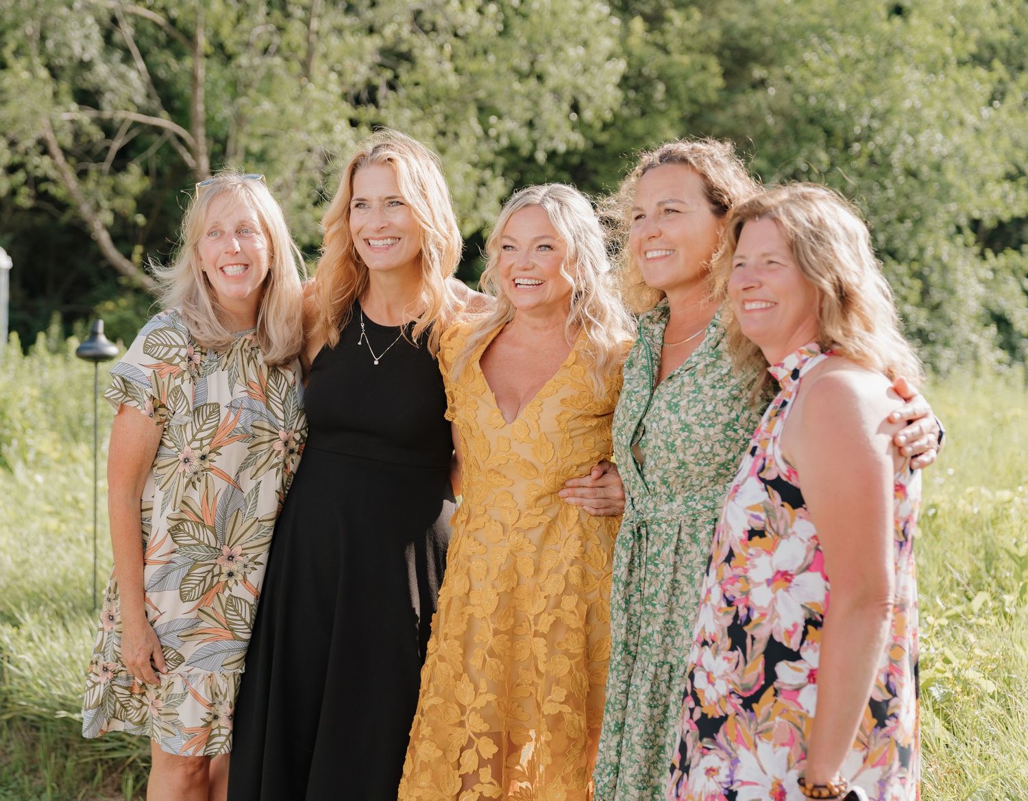 A group of five young women in colorful floral and solid dresses stand together in a natural outdoor setting.