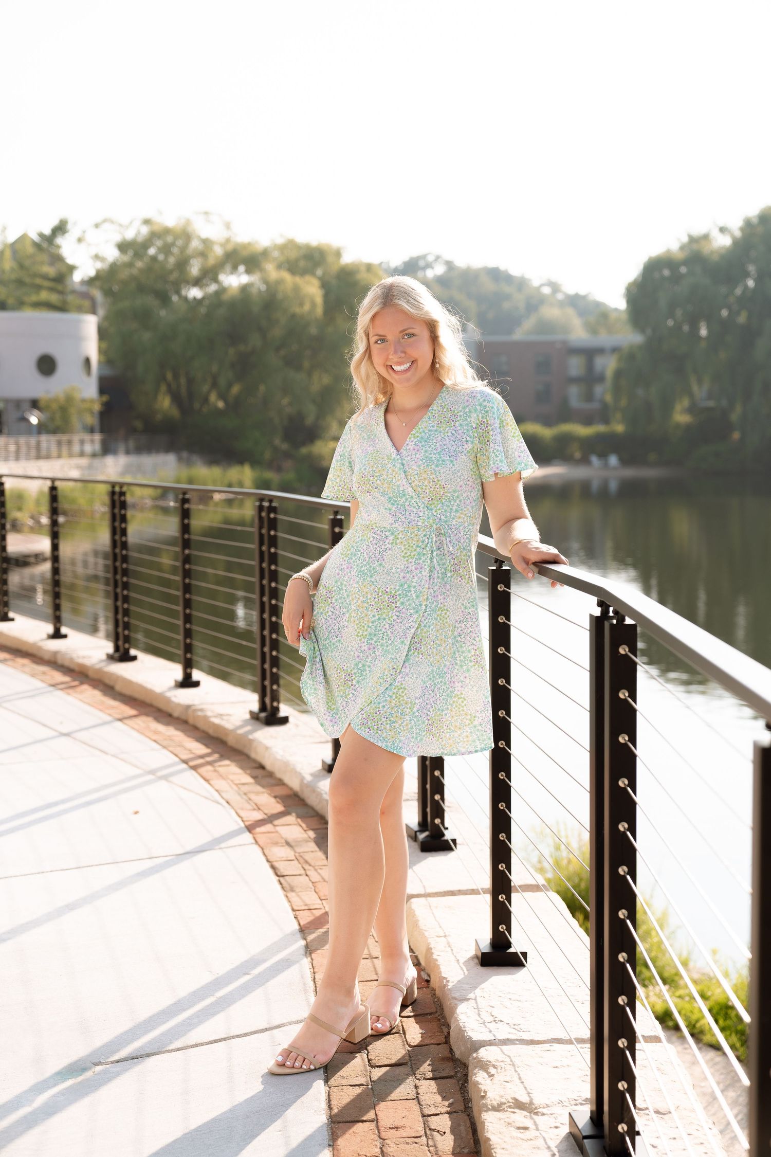 A smiling woman in a floral dress standing on a wooden walkway by a lake.