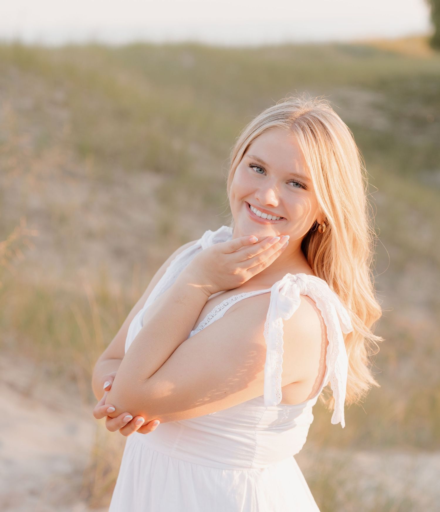 A smiling woman in a white dress stands in a field on a sunny day.