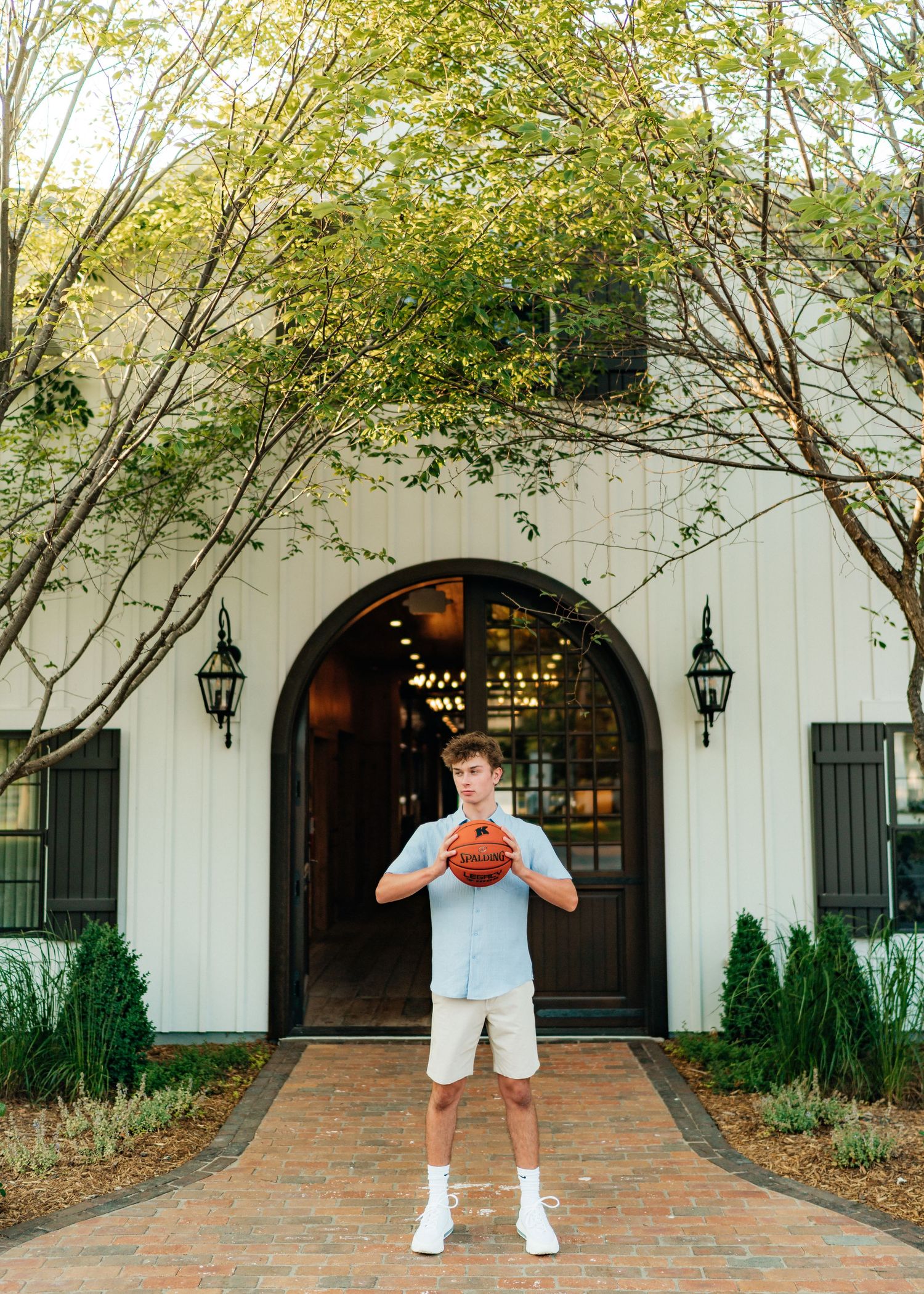 Teen boy holding basketball in front of elegant white arched building entrance with lush green trees.