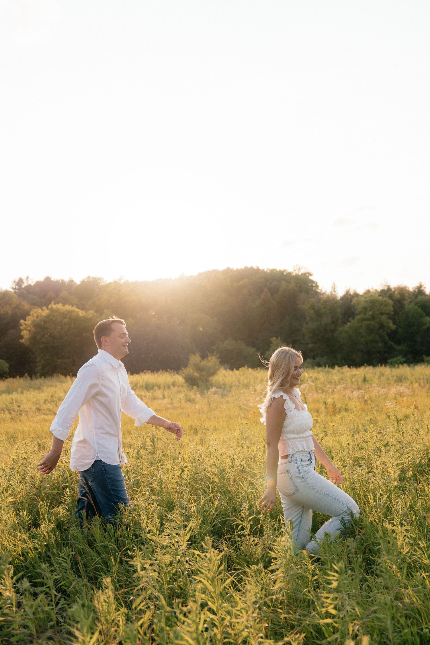 Couple walking through a sunny golden field at sunset, man in white shirt and woman in jeans enjoying a romantic outdoor stroll.