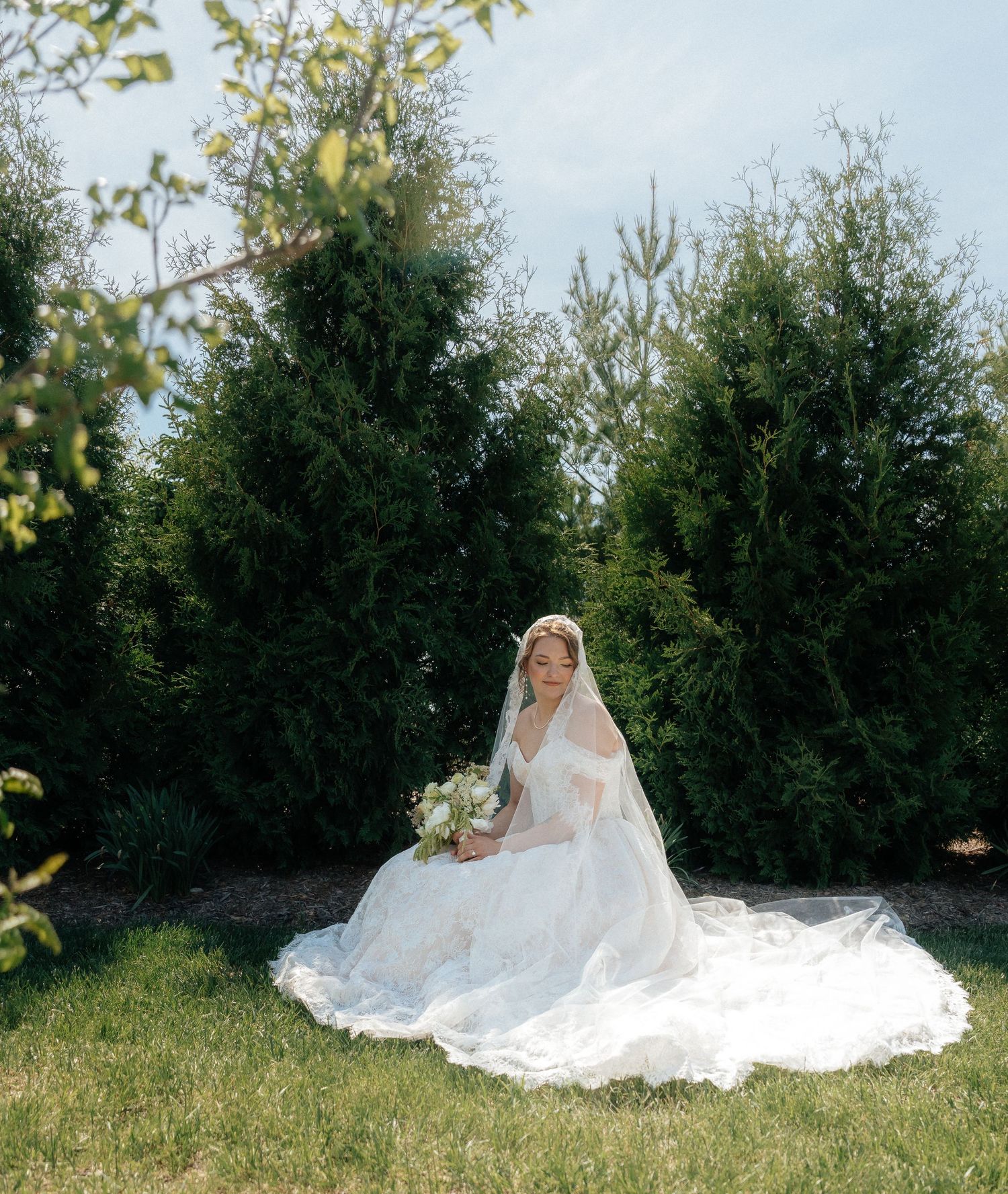 Bride in white gown and veil sits on grass holding bouquet, surrounded by tall evergreen trees outdoors.