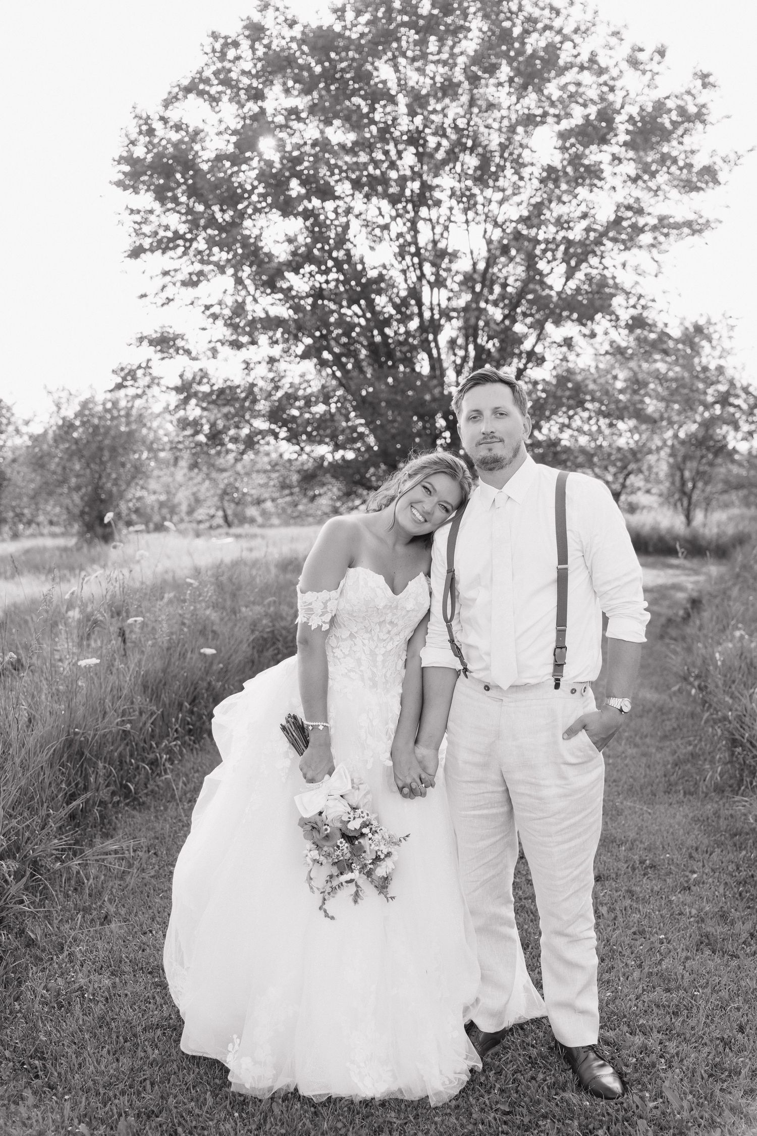 A black and white photograph of a bride and groom standing in a field with a tree in the background.