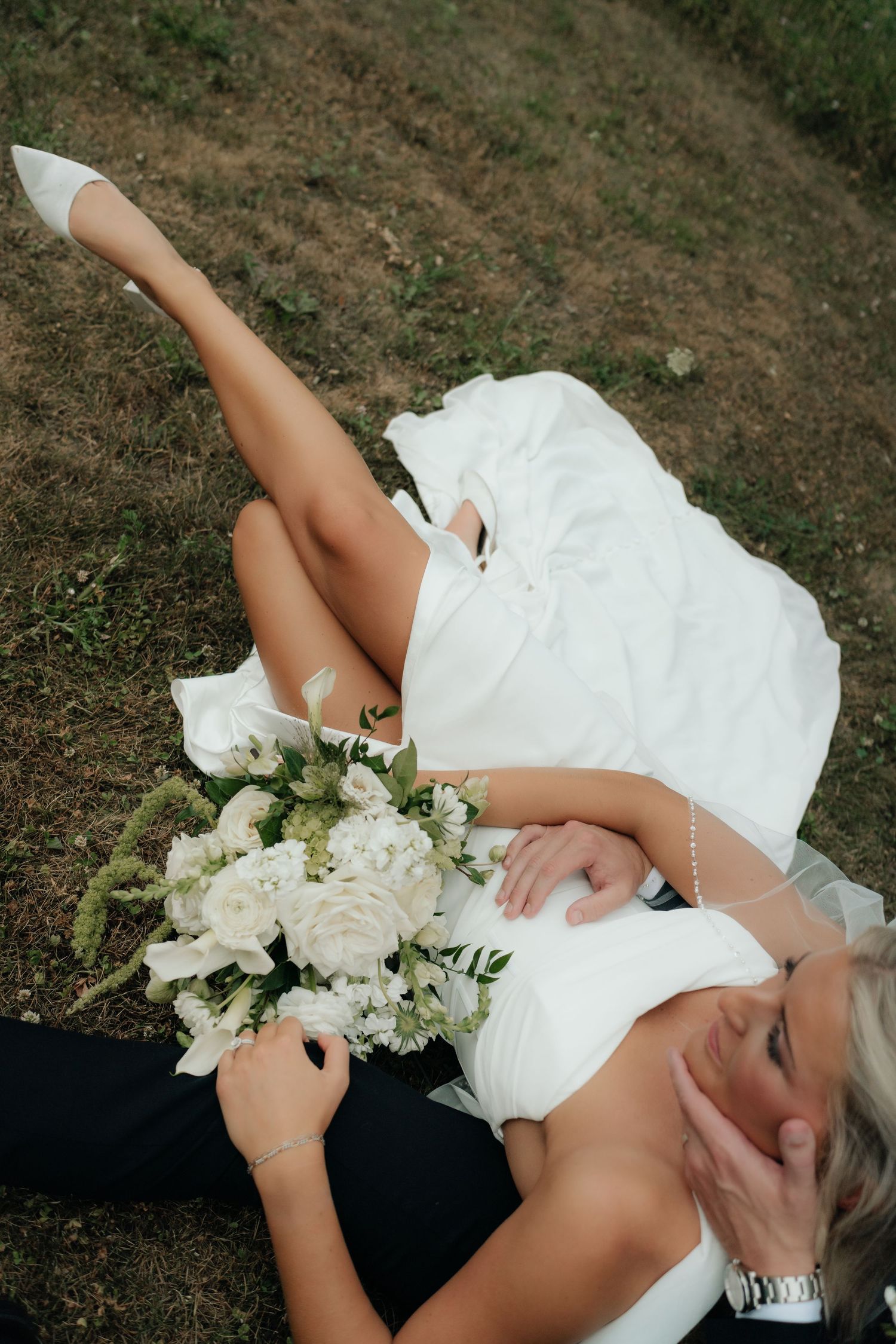 Bride in white wedding dress lying on grass with bouquet of white flowers, legs raised, smiling joyfully.