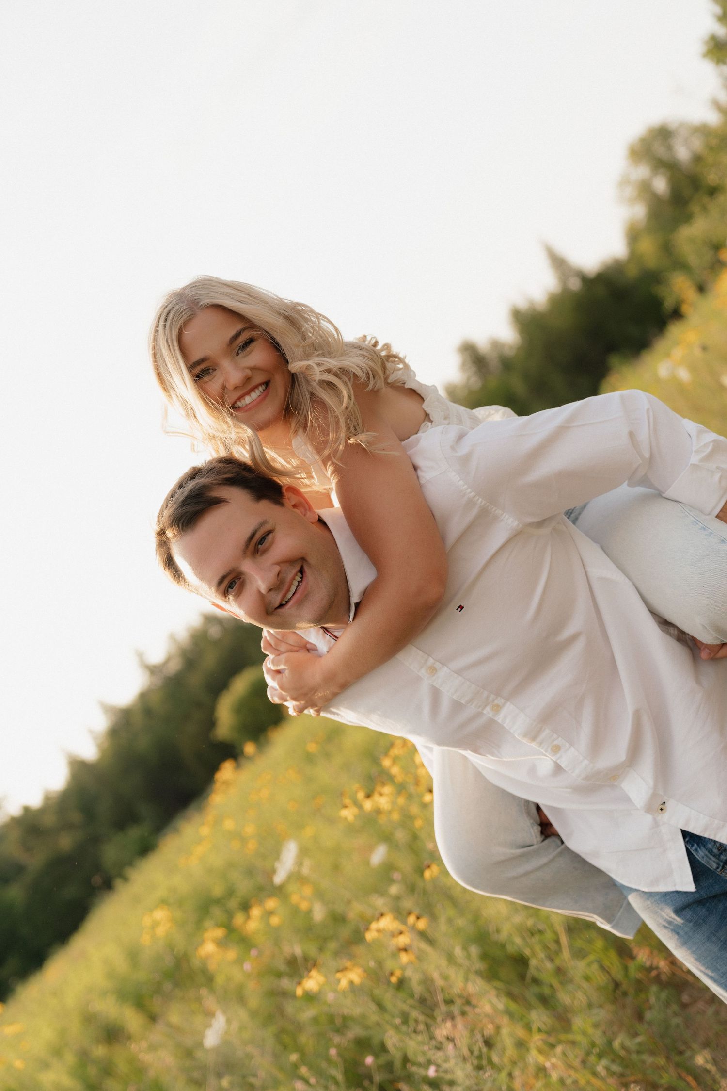 Happy couple in white outfits enjoying a piggyback ride in a sunny green field with wildflowers.