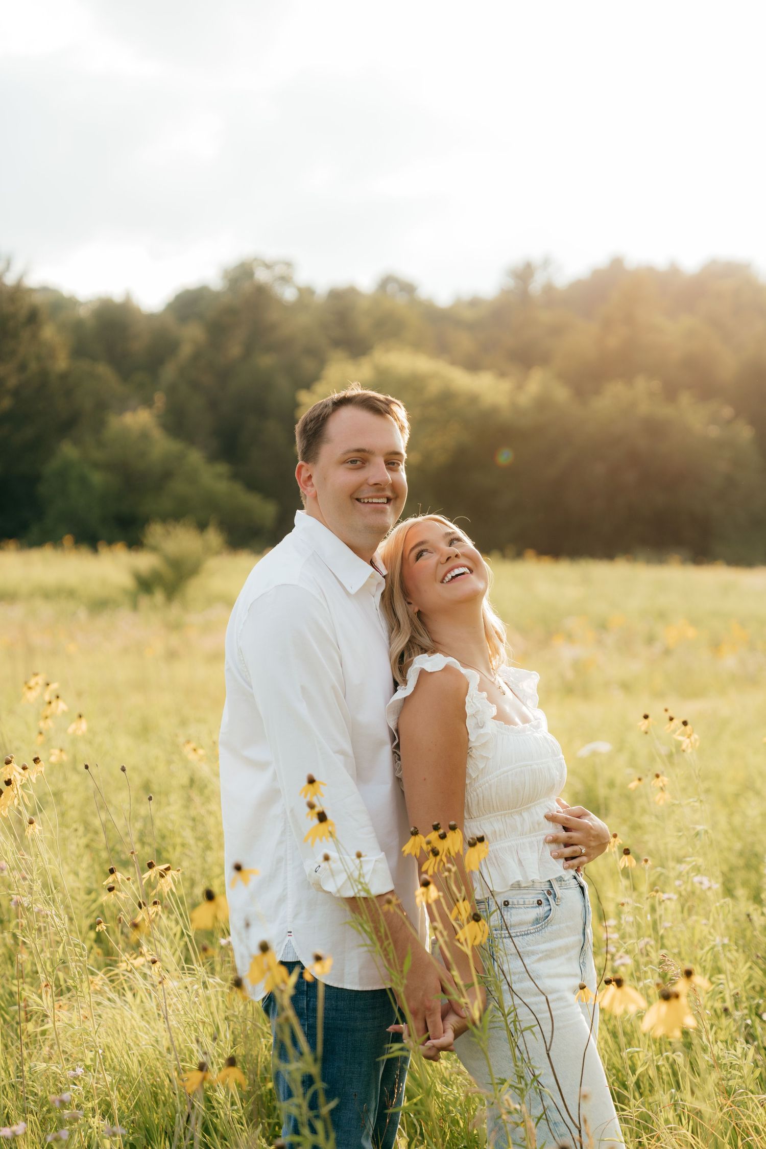 A happy couple embraces in a sunny wildflower field, the woman laughing joyfully while the man smiles warmly behind her.