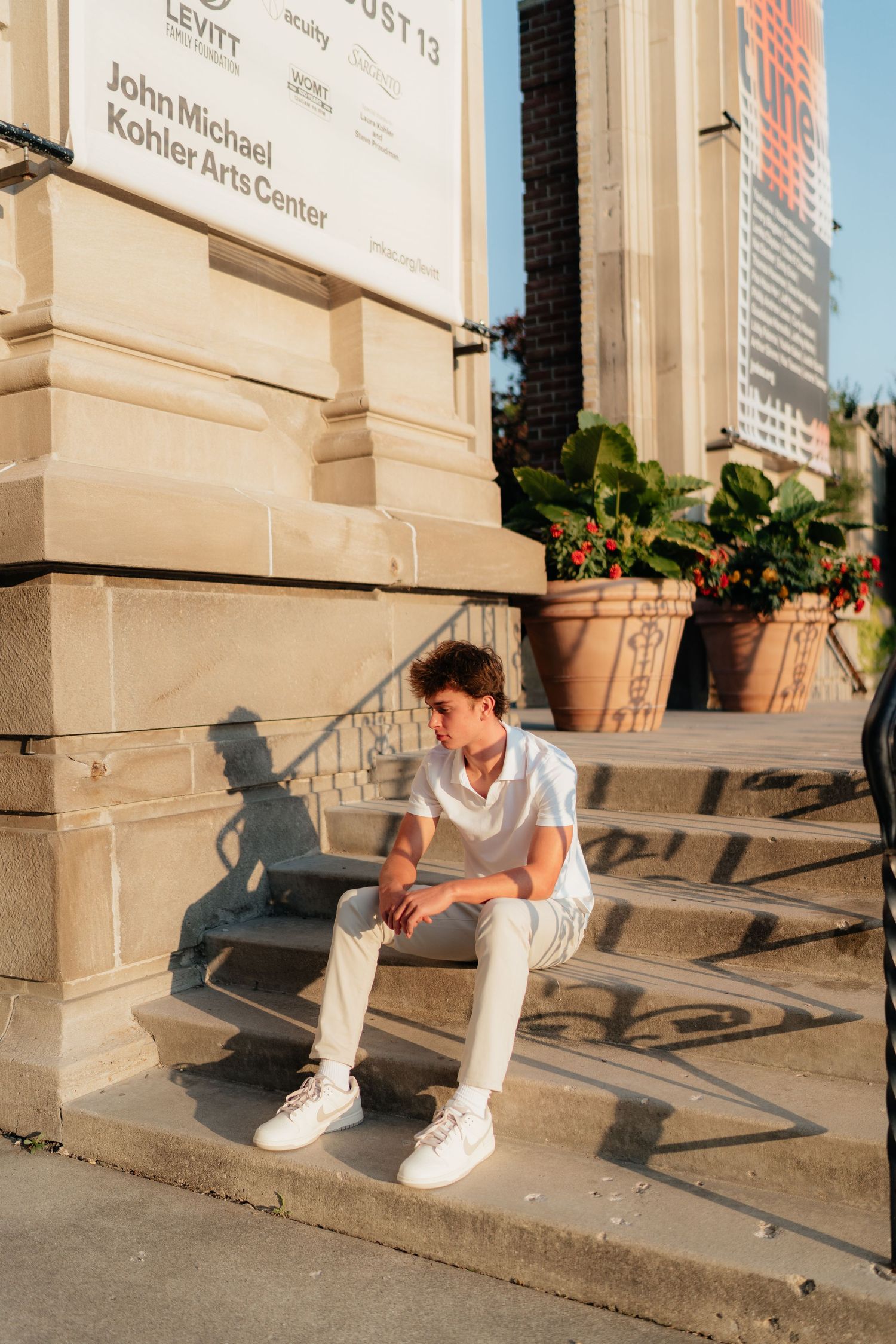 Young man in white outfit sitting on steps outside John Michael Kohler Arts Center in golden hour light.