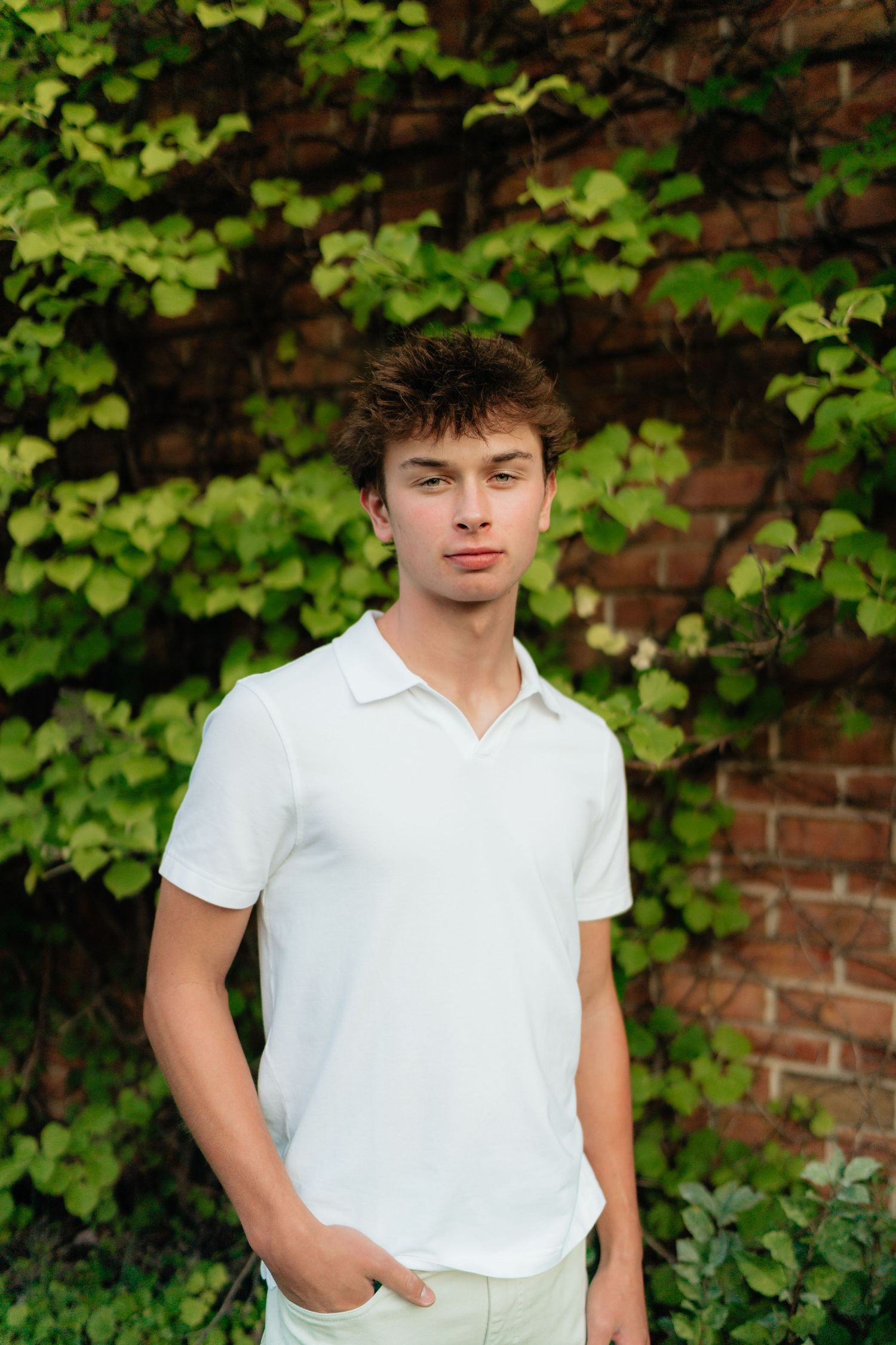 Young man in white polo shirt standing outdoors against brick wall covered in green ivy leaves.