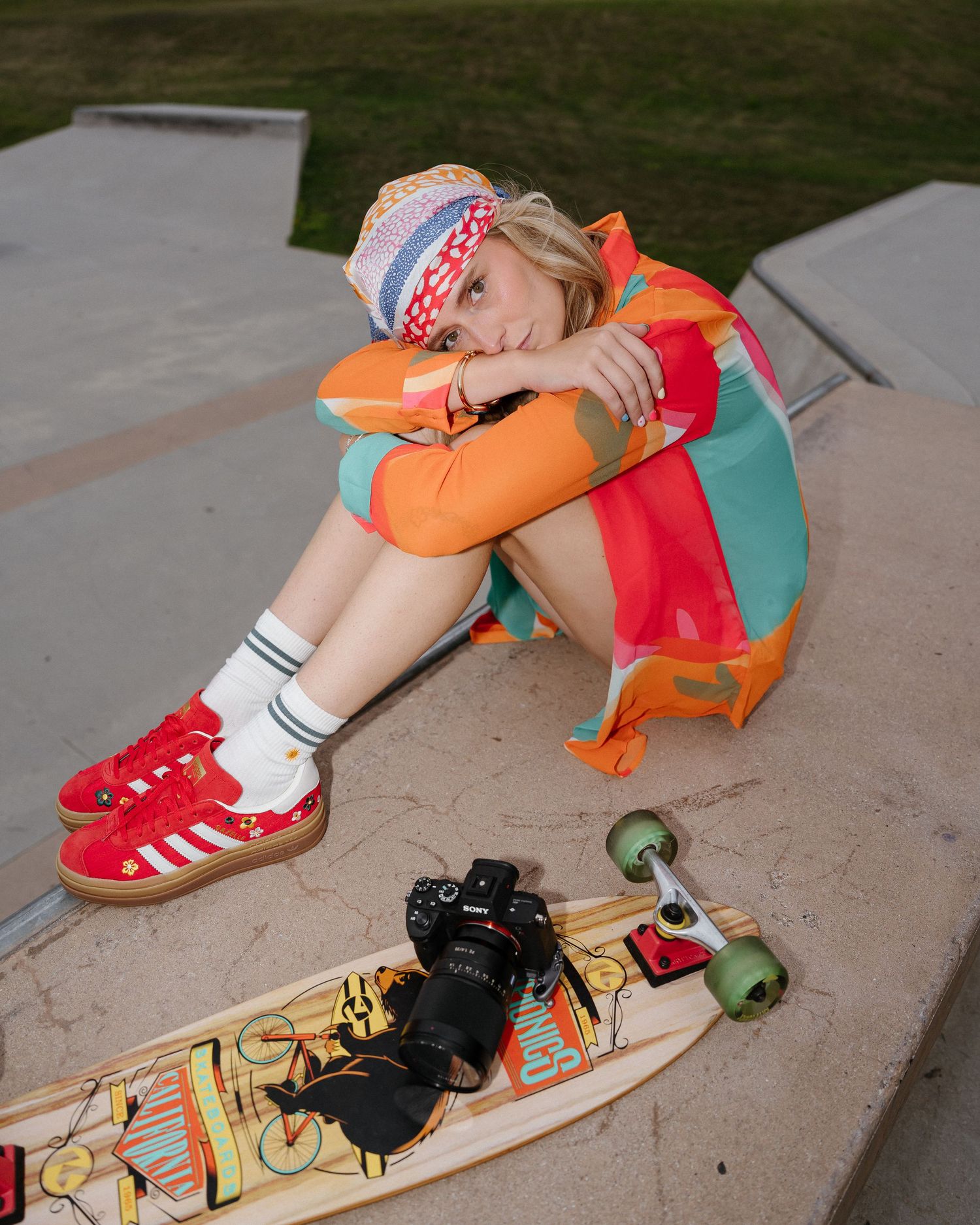 Young woman in colorful outfit and red Adidas sneakers sits at skate park with longboard and camera nearby.