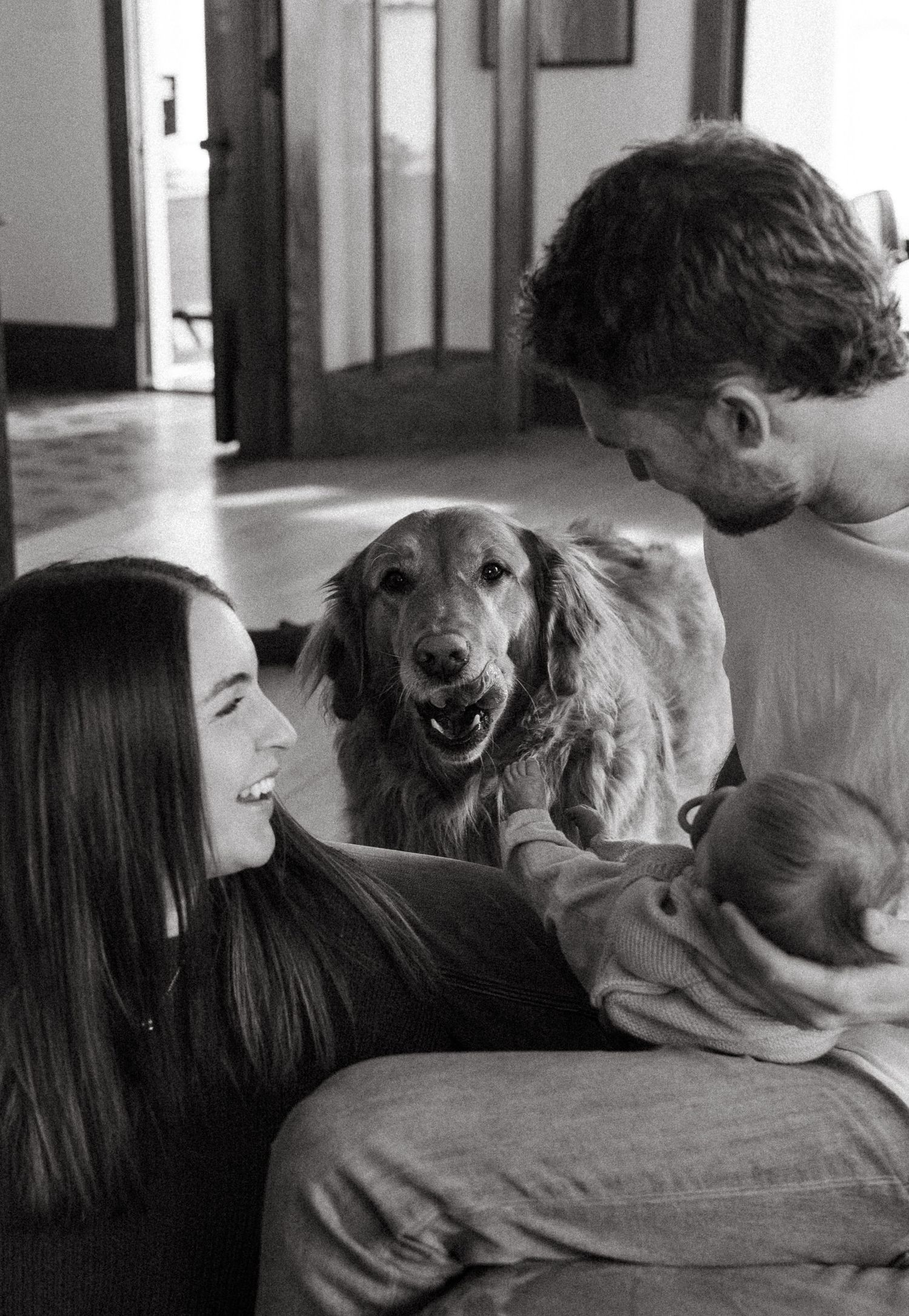 A black and white image shows a family with a dog sitting on a couch, with a smiling woman holding a baby.