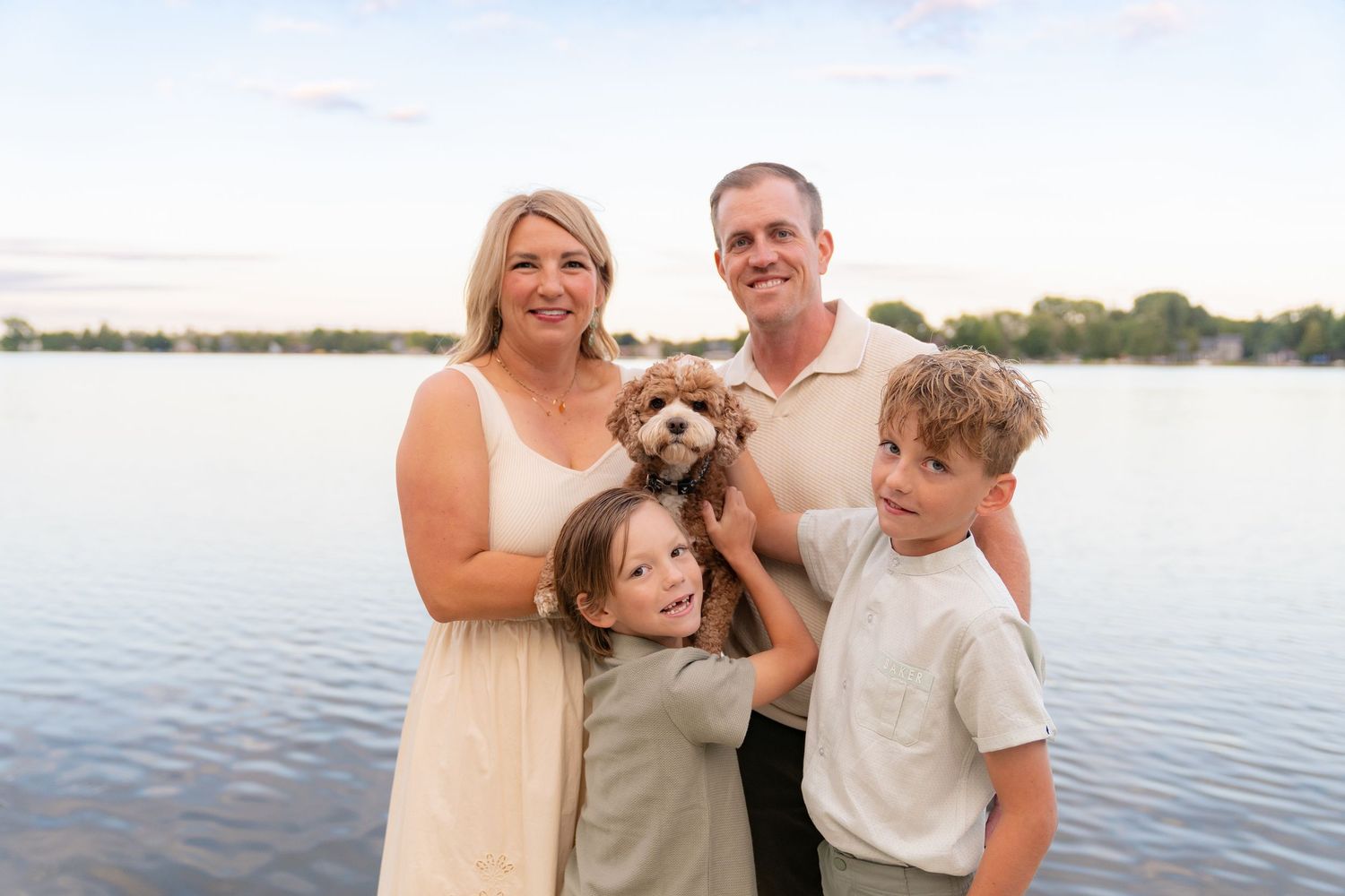 A happy family of four, including a couple and two children, posing together with a small dog by a lake.