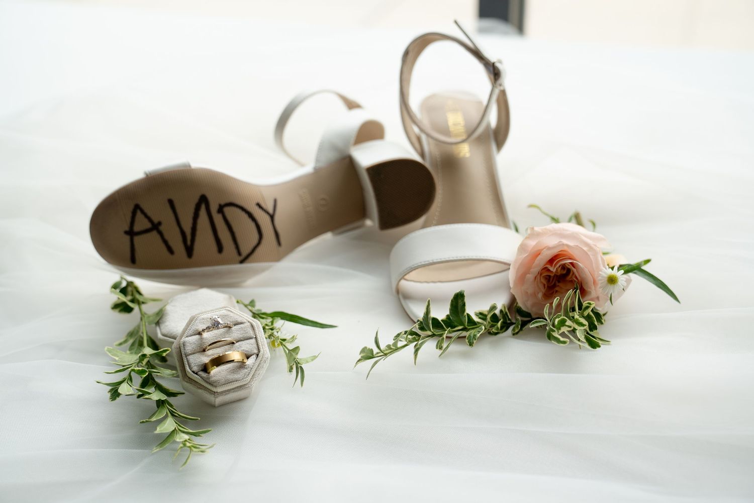 A pair of white and tan wedding shoes with the name 'ANDY' written on them, surrounded by greenery.