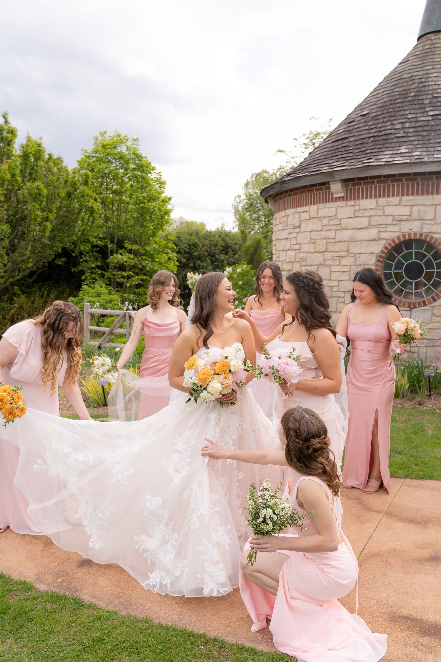 A bride and her bridesmaids in pink dresses pose together in a garden setting with a stone building in the background.
