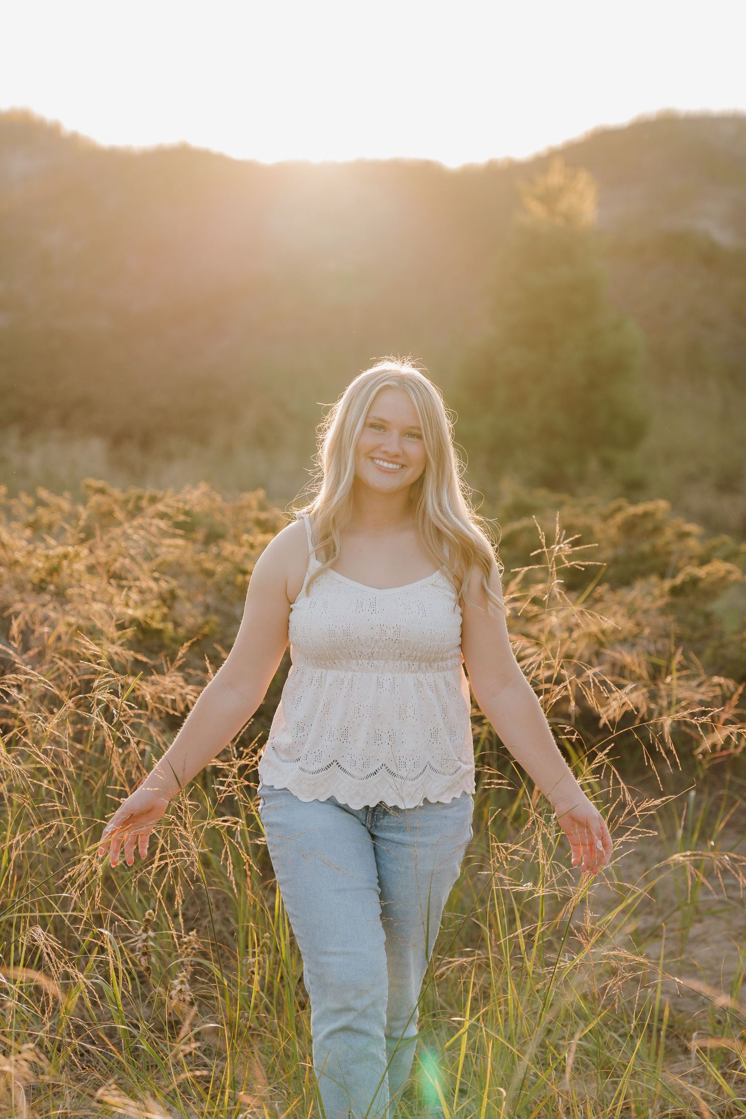 A young woman in a white top and blue jeans walks through a field of tall grass at sunset.
