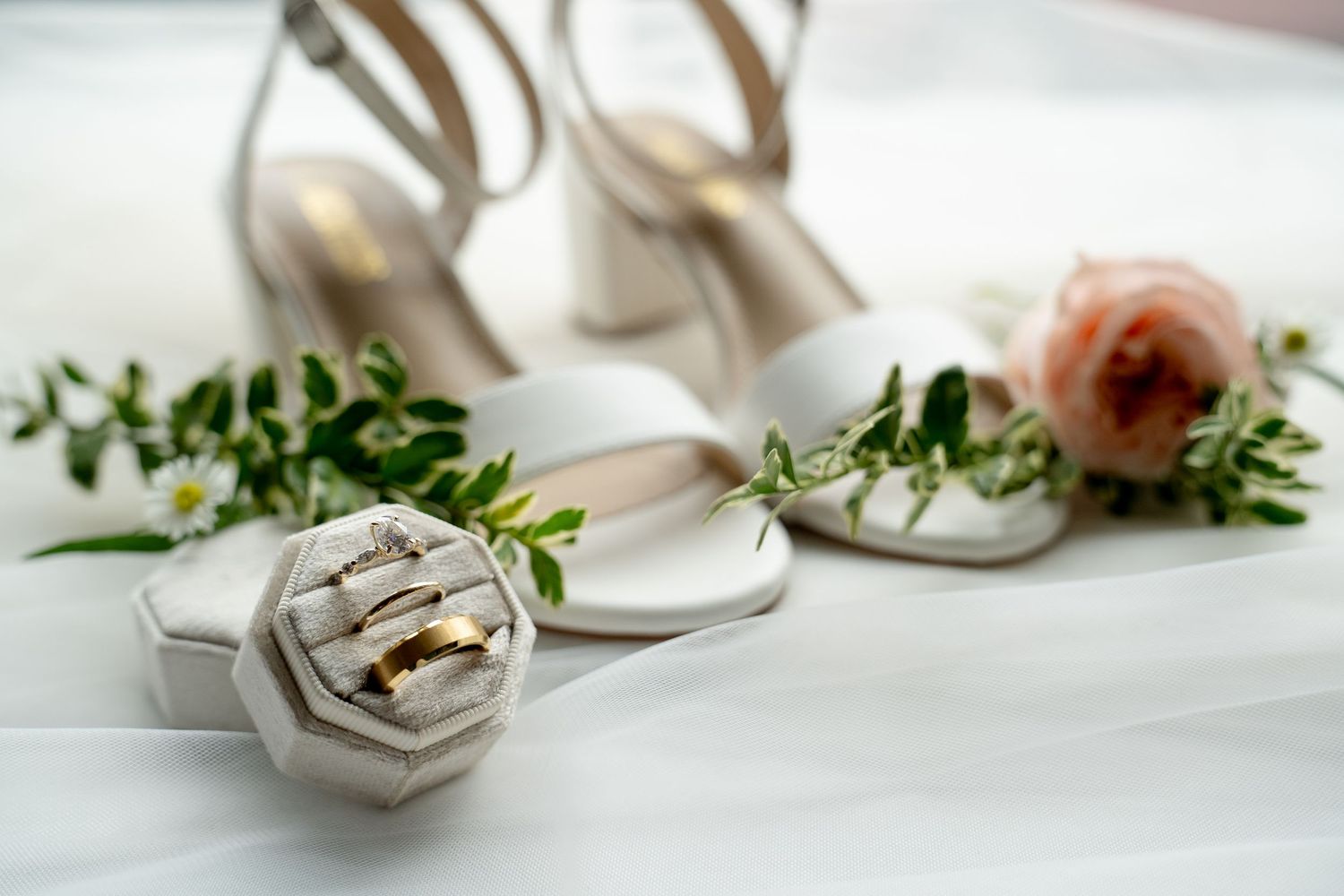 The image shows a pair of white high-heeled shoes, a green plant, and a ring on a white surface.