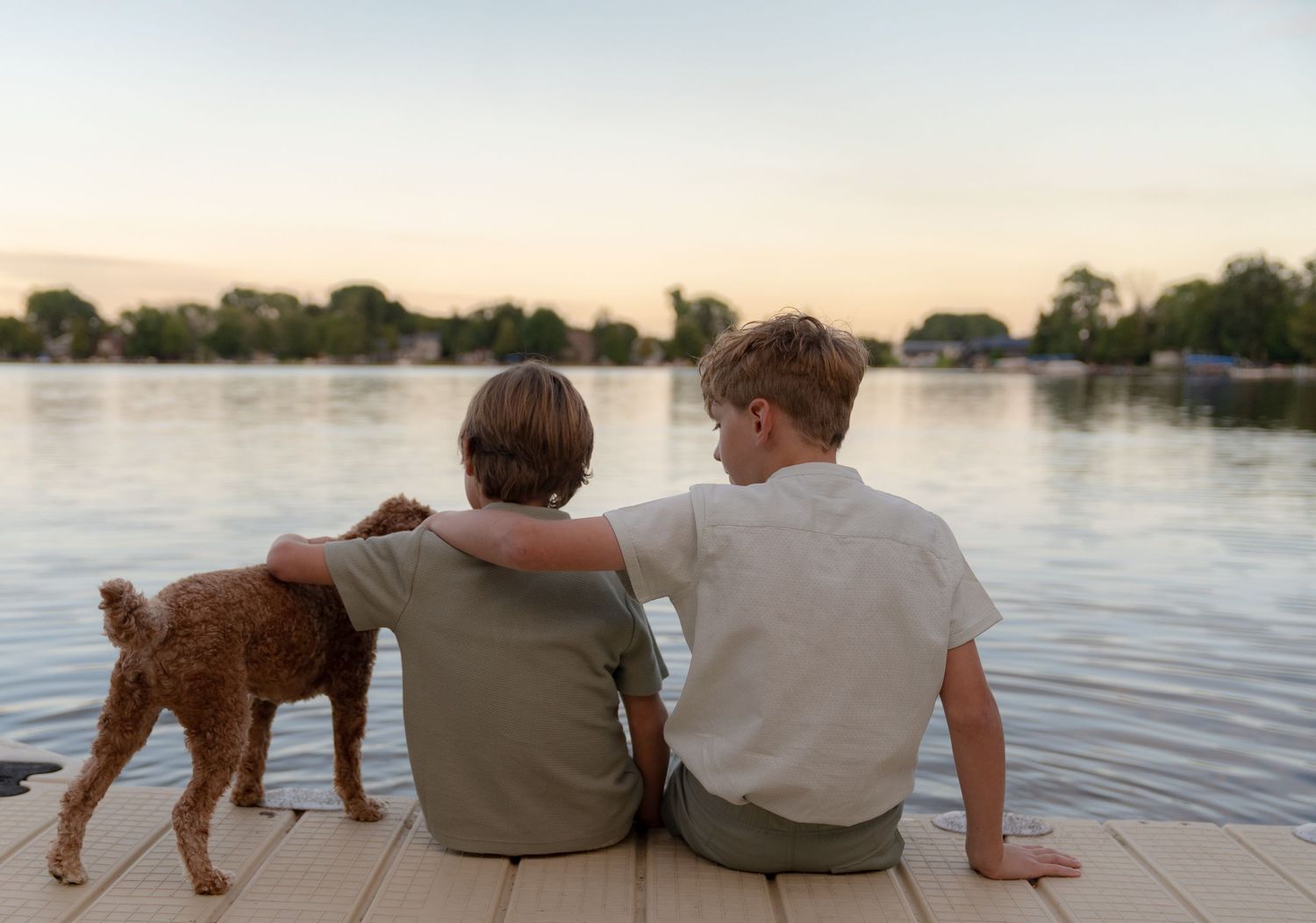 Two young boys sitting on a riverbank, petting a small dog while enjoying the peaceful scenery.