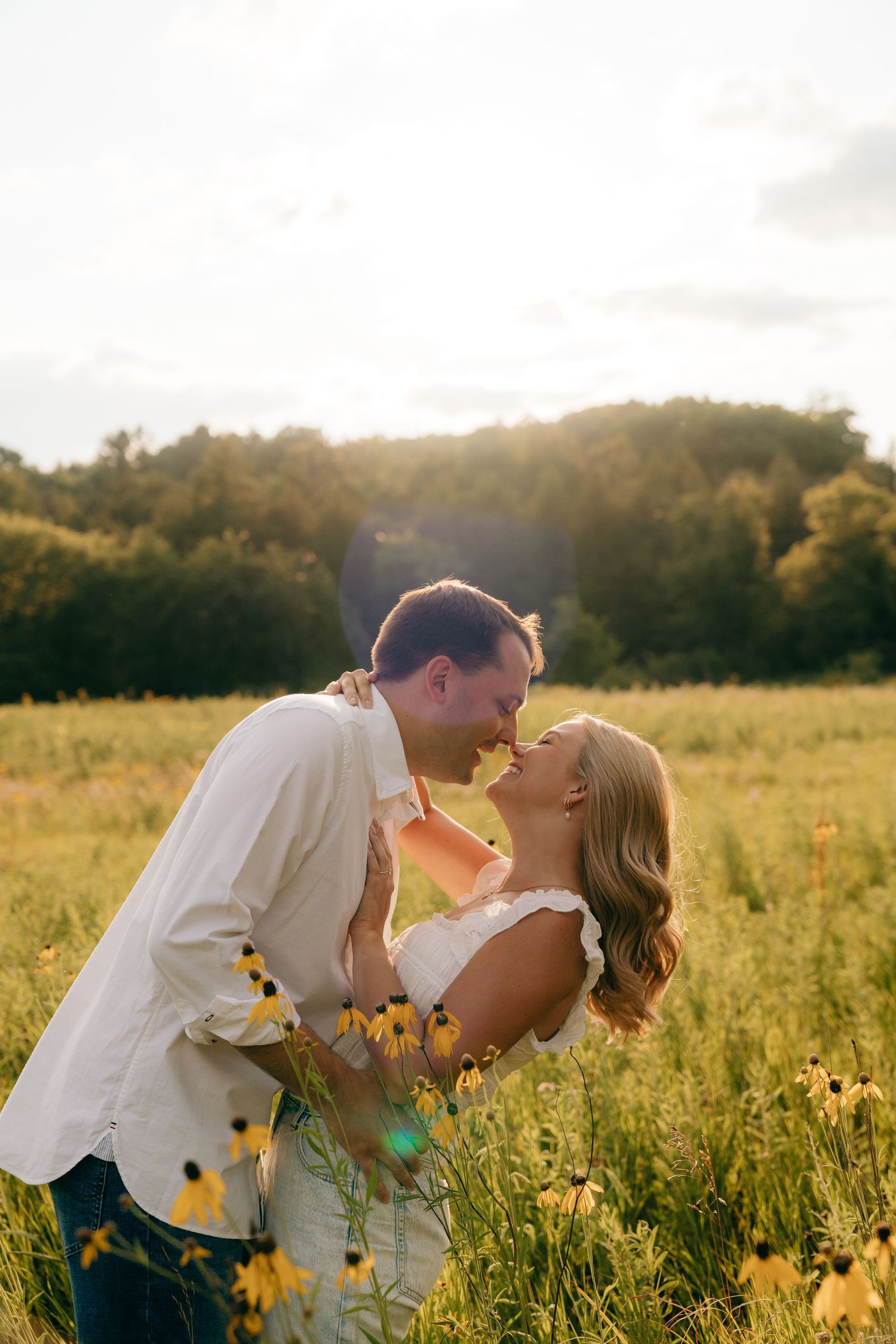 Couple sharing a romantic kiss in a sunlit wildflower field with yellow blooms and lush green trees in the background.