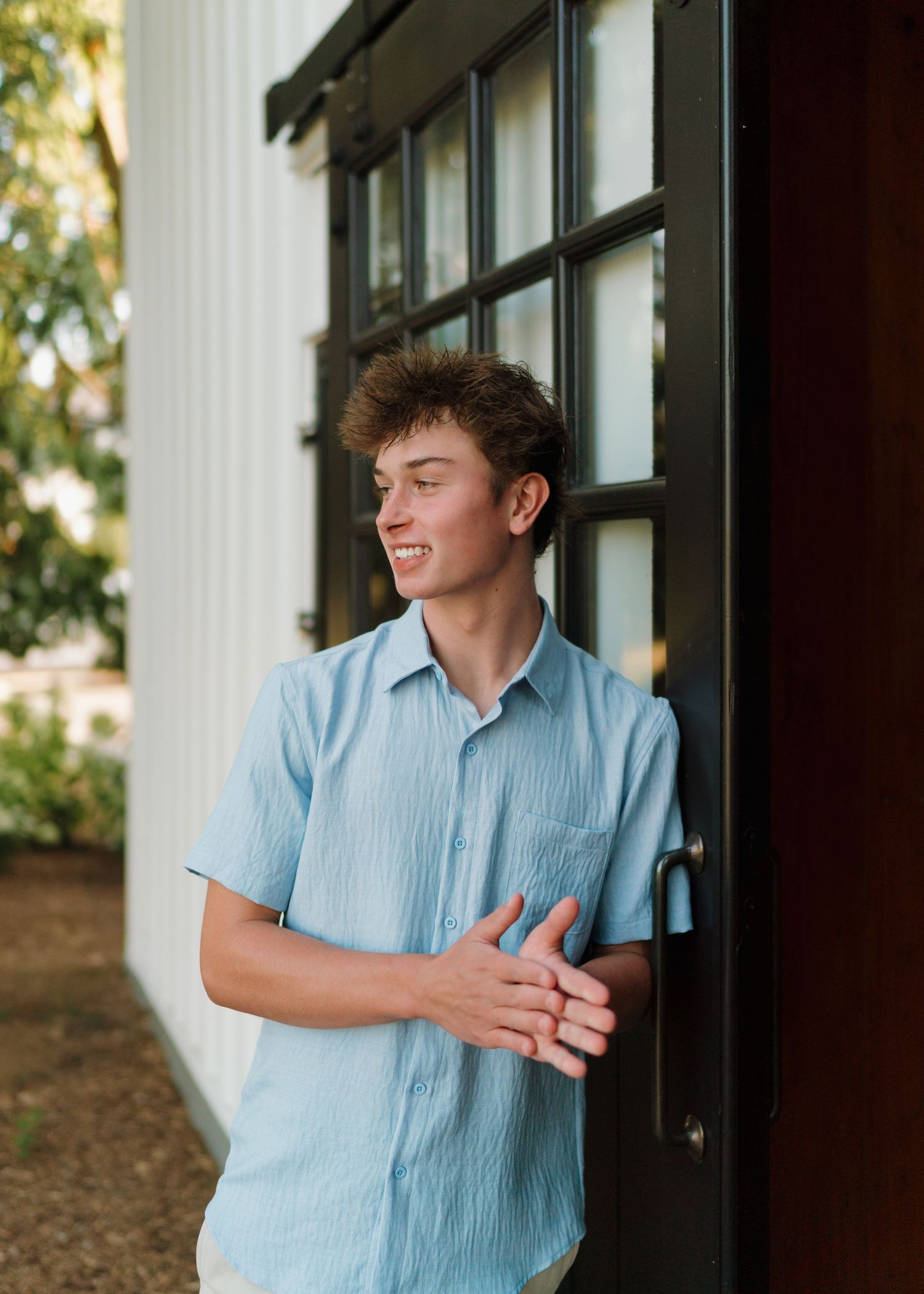 Smiling teenage boy in light blue short-sleeve shirt leaning against a dark door frame outdoors.