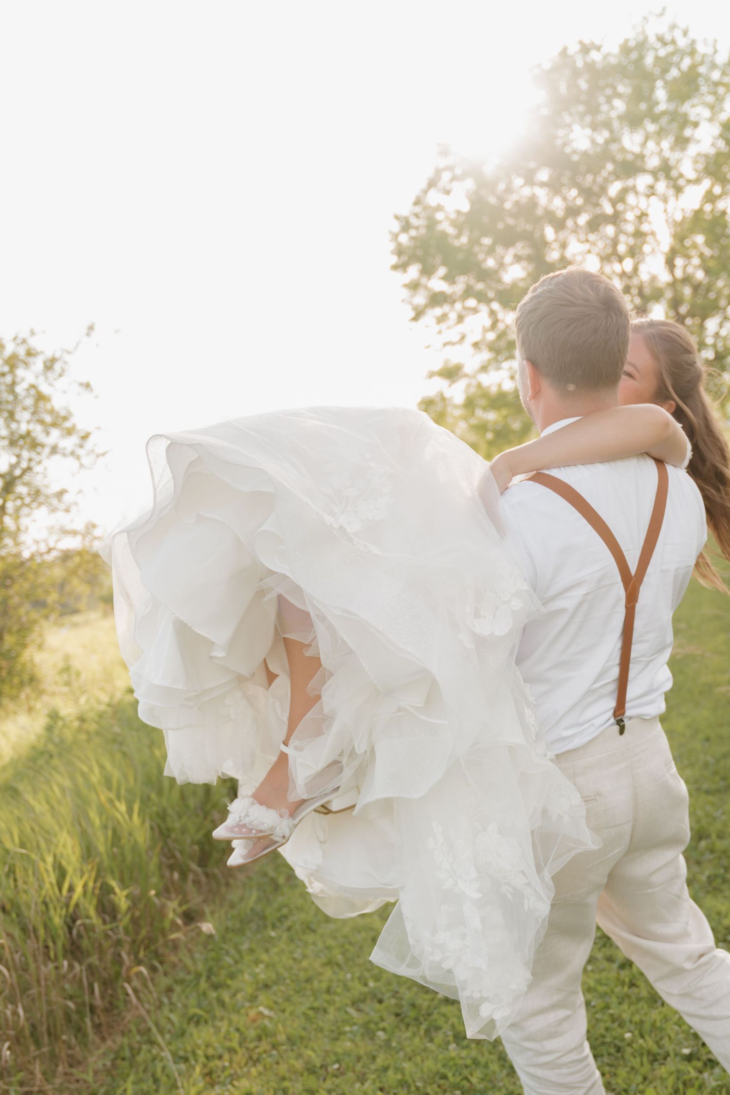A bride in a flowing white dress is being carried by her groom in a grassy field with trees in the background.
