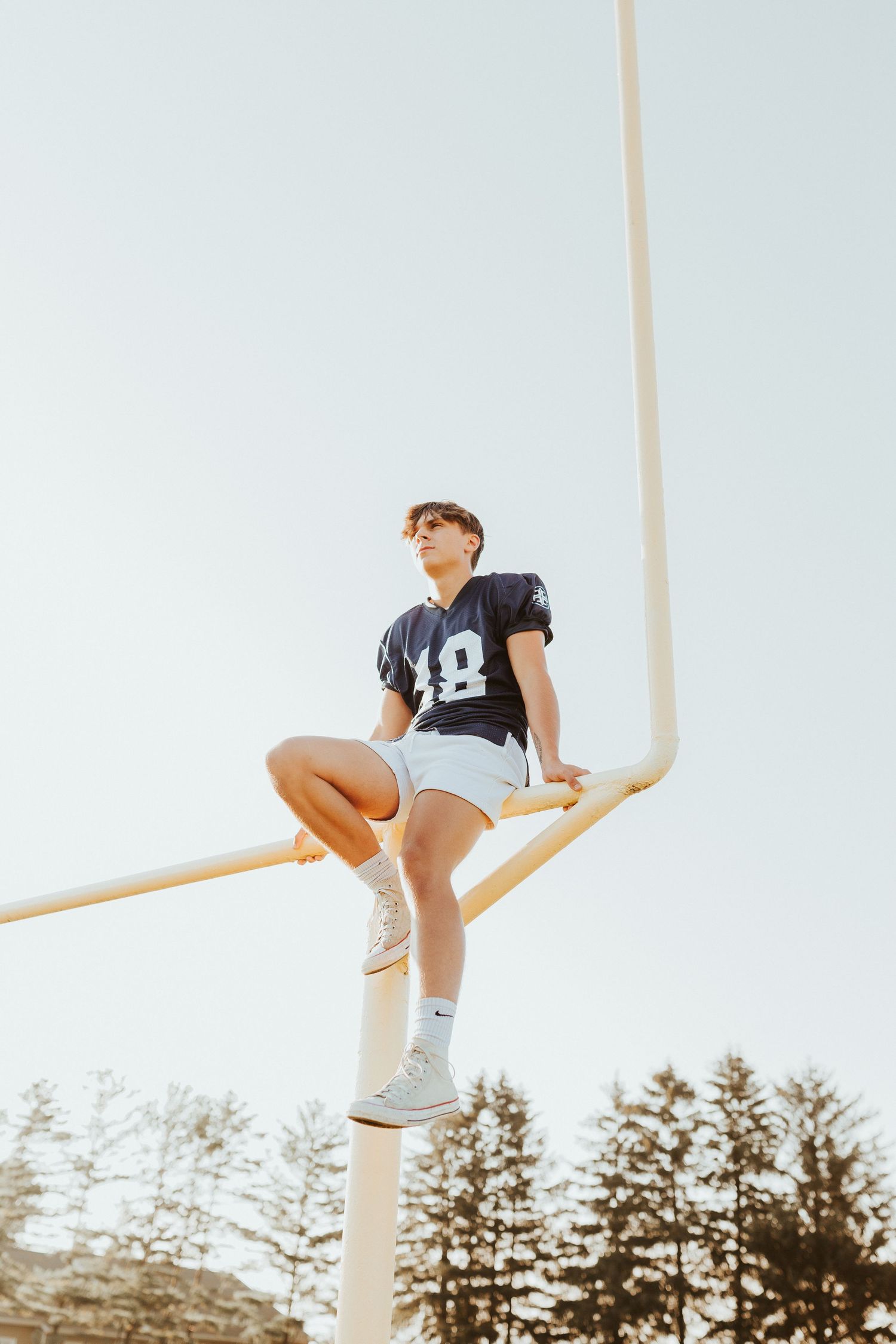 A young person wearing a sports jersey is sitting on a wooden beam against a background of trees.