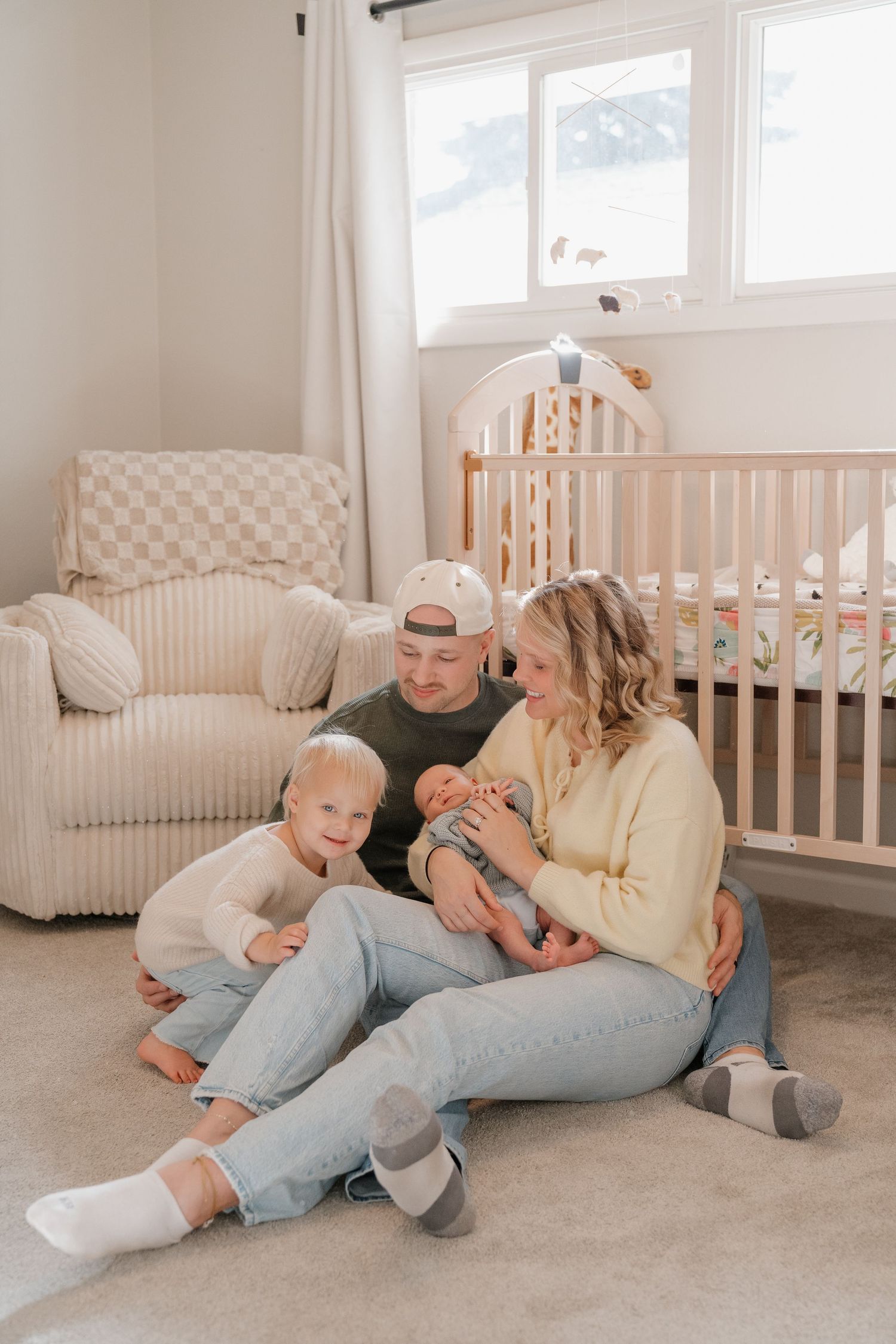 A happy family of three sits on the floor in a cozy nursery with a crib and comfortable chair.