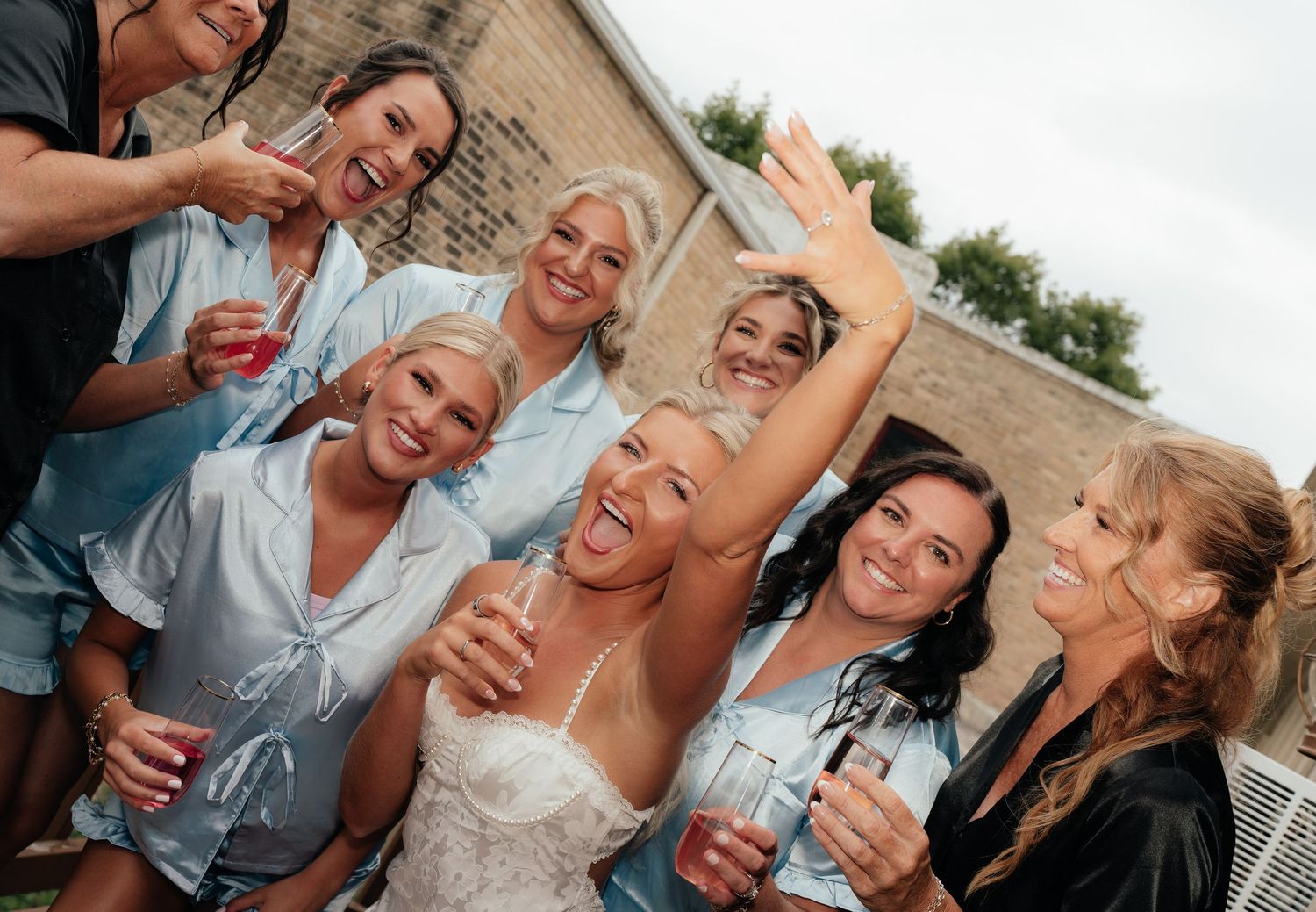 Bride in white lace gown celebrates with bridesmaids in blue robes, holding drinks and smiling outdoors.