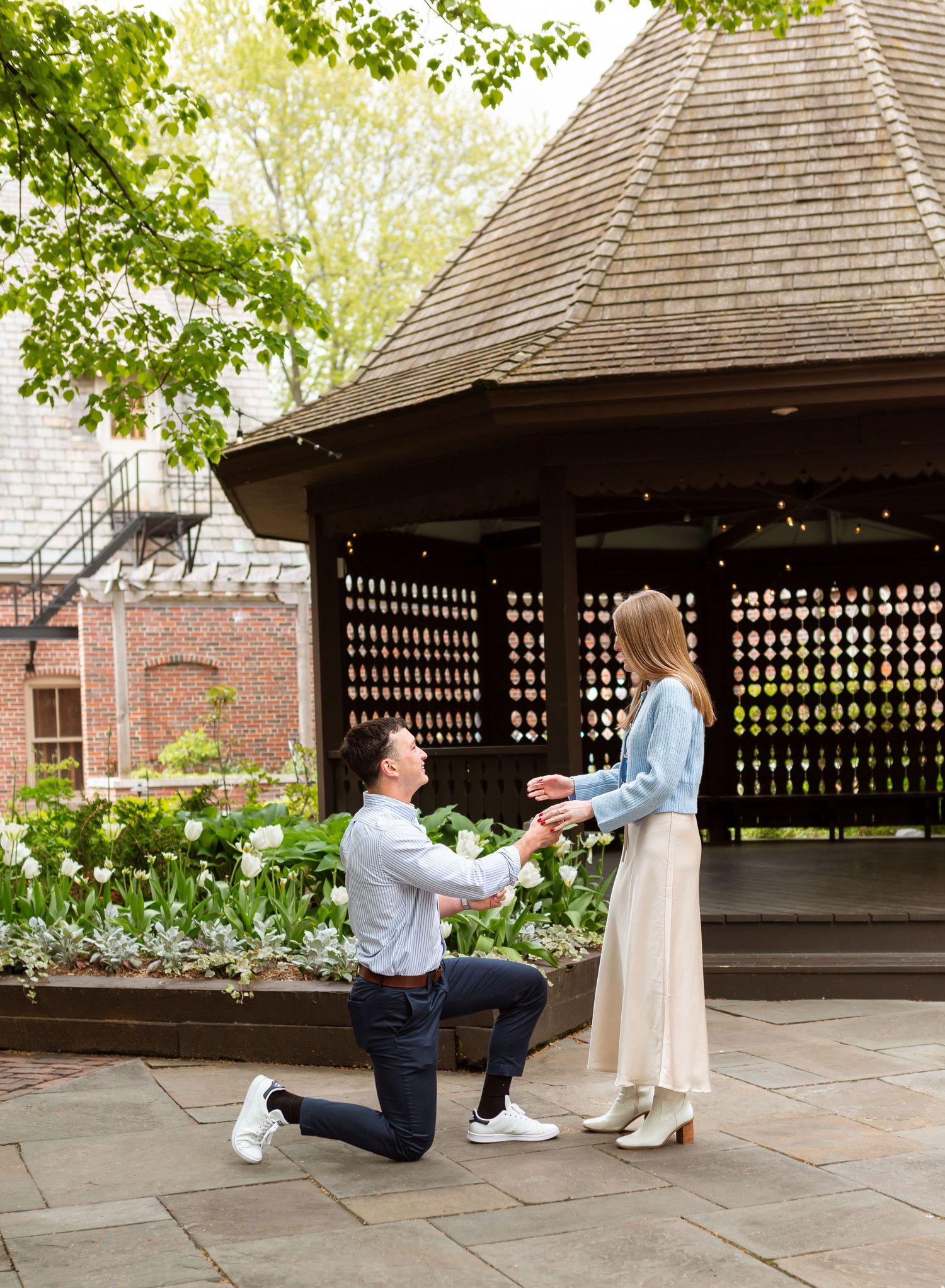 A man kneeling and proposing to a woman in a garden with a gazebo in the background.