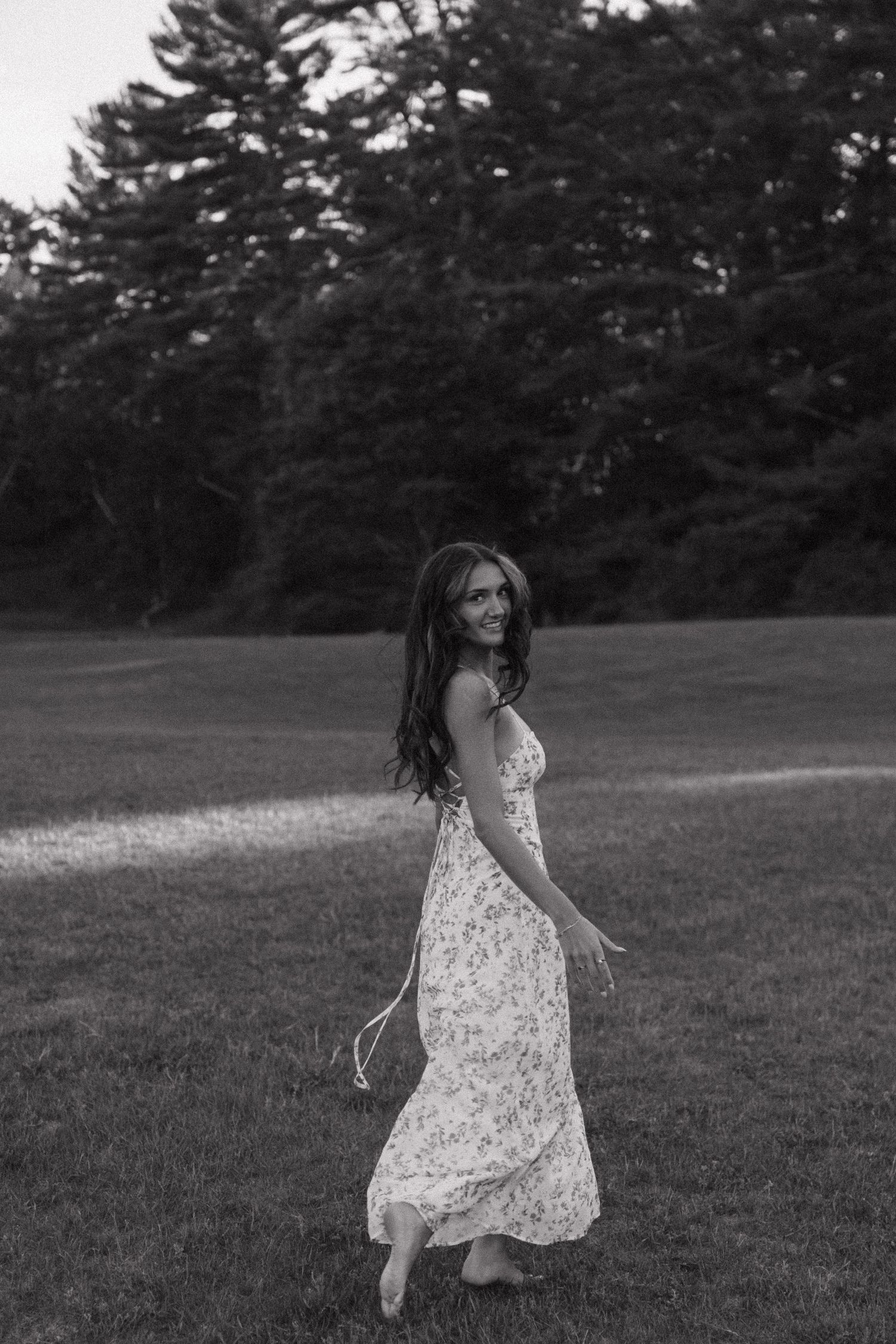 Black and white photo of a smiling woman in a floral maxi dress walking barefoot across a lush green lawn.