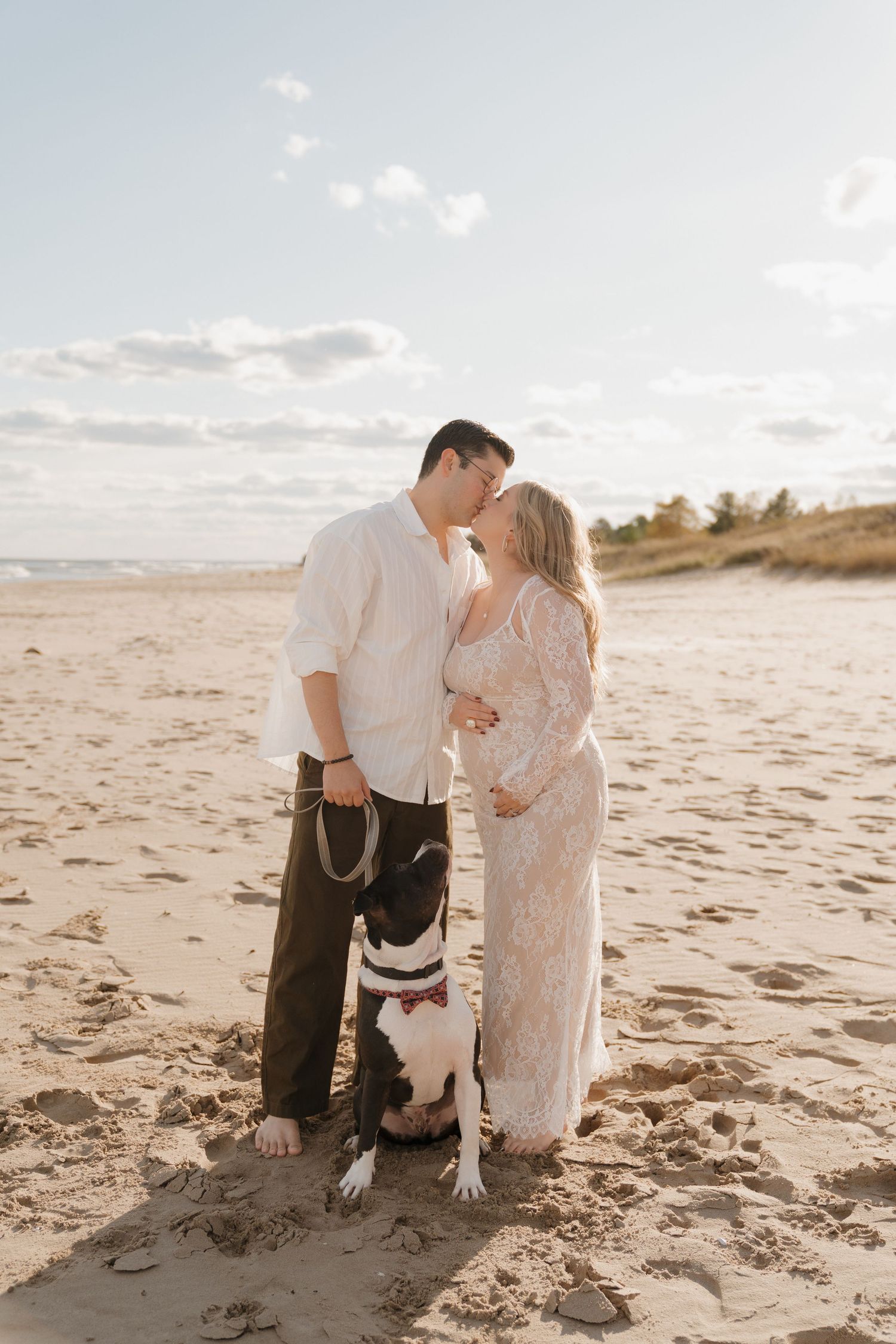 Couple kissing on beach with dog at golden hour, woman in white lace maternity dress.