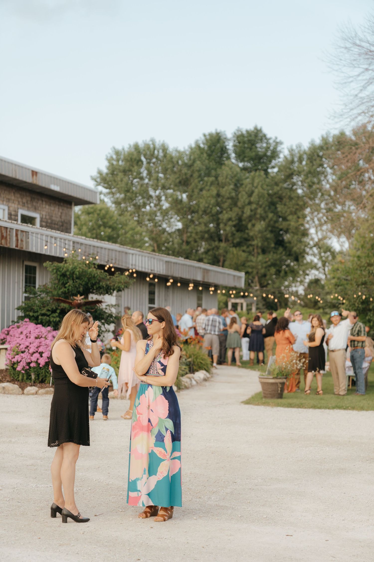A group of people gathered outdoors at a rustic event venue with string lights and trees in the background.