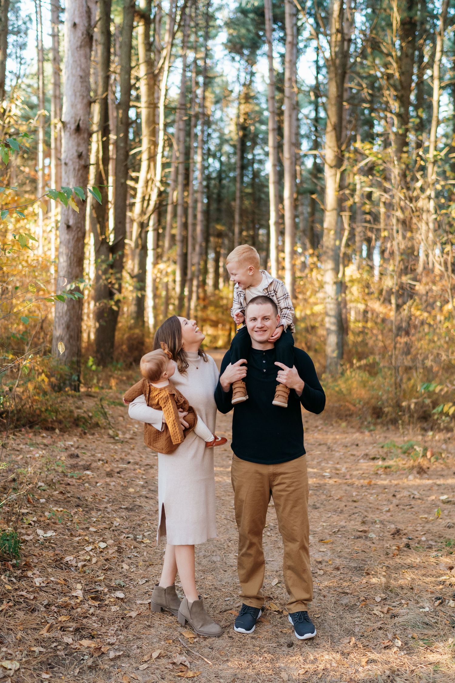 Family of four poses in autumn forest, parents holding young children among golden foliage and tall pine trees.