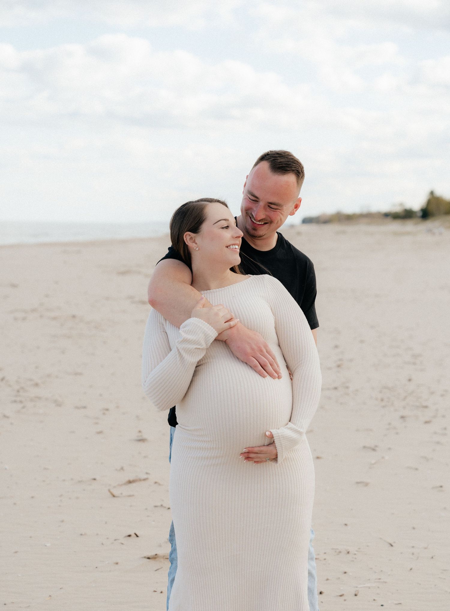 Pregnant couple embracing on a sandy beach, woman in white ribbed dress, man in black shirt, smiling together.