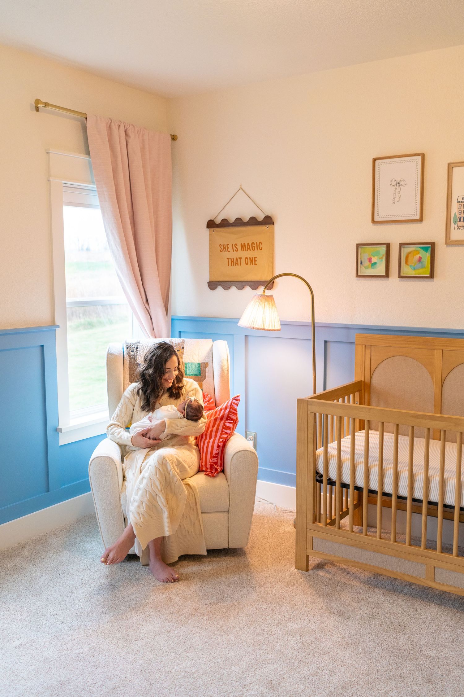 A woman sits in a rocking chair in a cozy nursery, holding a baby and surrounded by decorative elements.