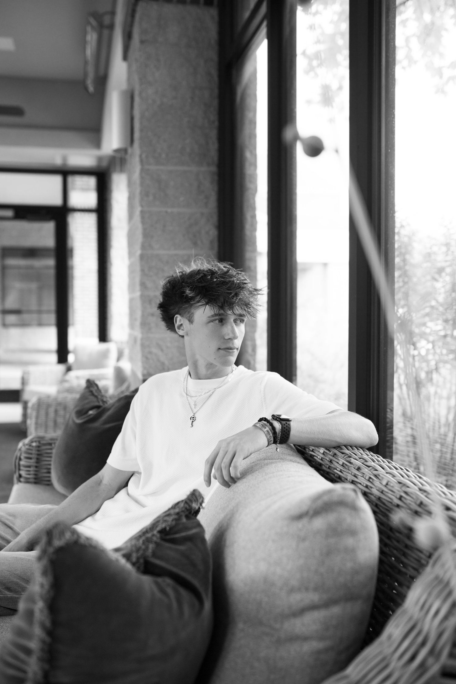 A young man with curly hair sits on a couch, looking thoughtfully out a window.