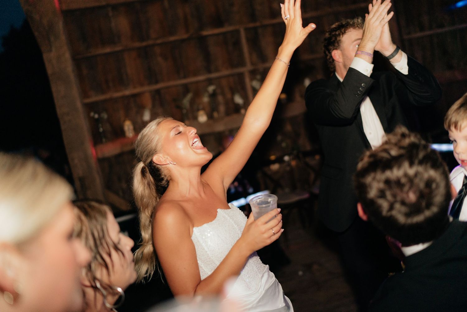 Bride in white dress dances with raised arms at lively indoor wedding reception celebration.