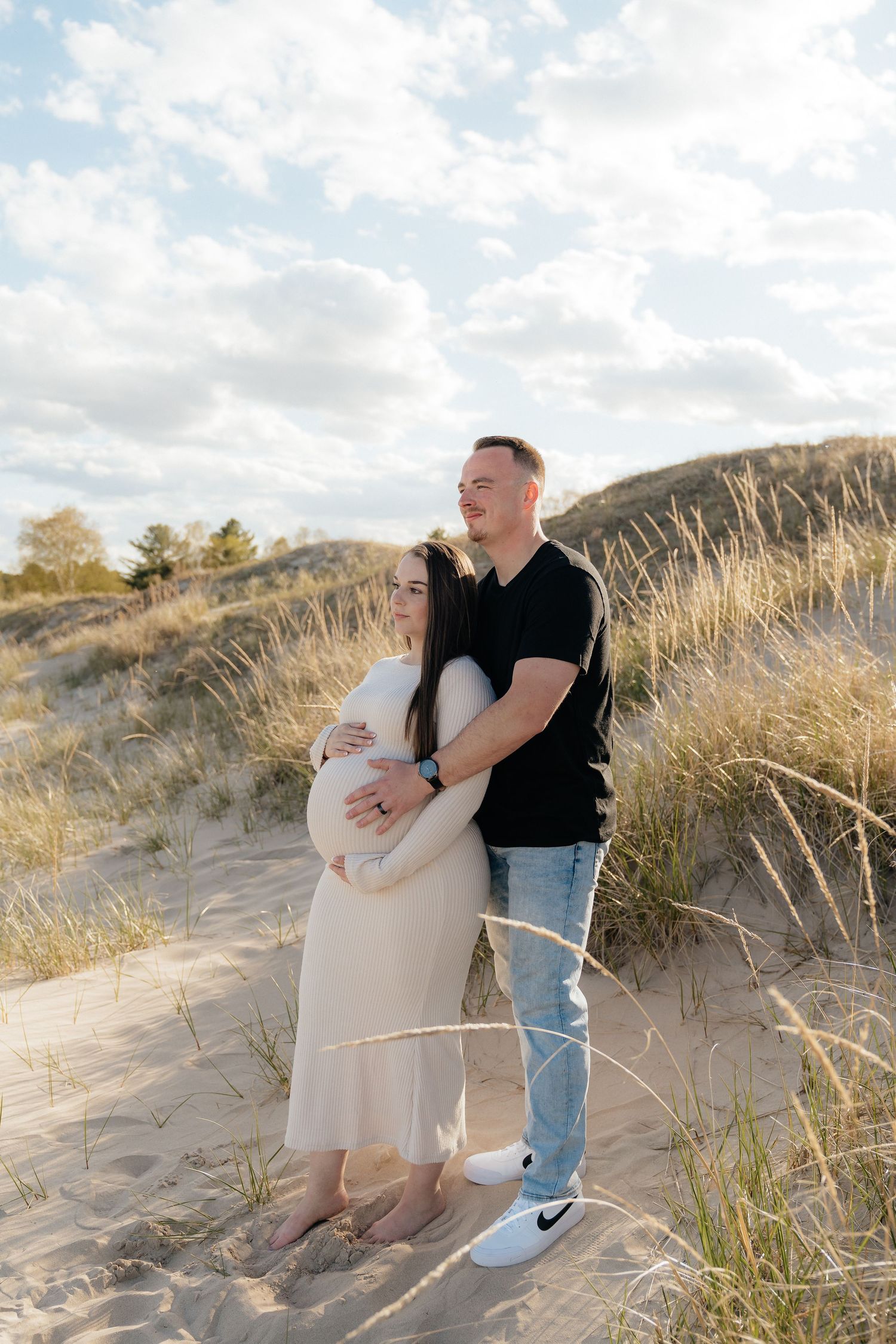 A couple stands on sandy dunes, the man embracing a pregnant woman in a white dress, surrounded by beach grass.