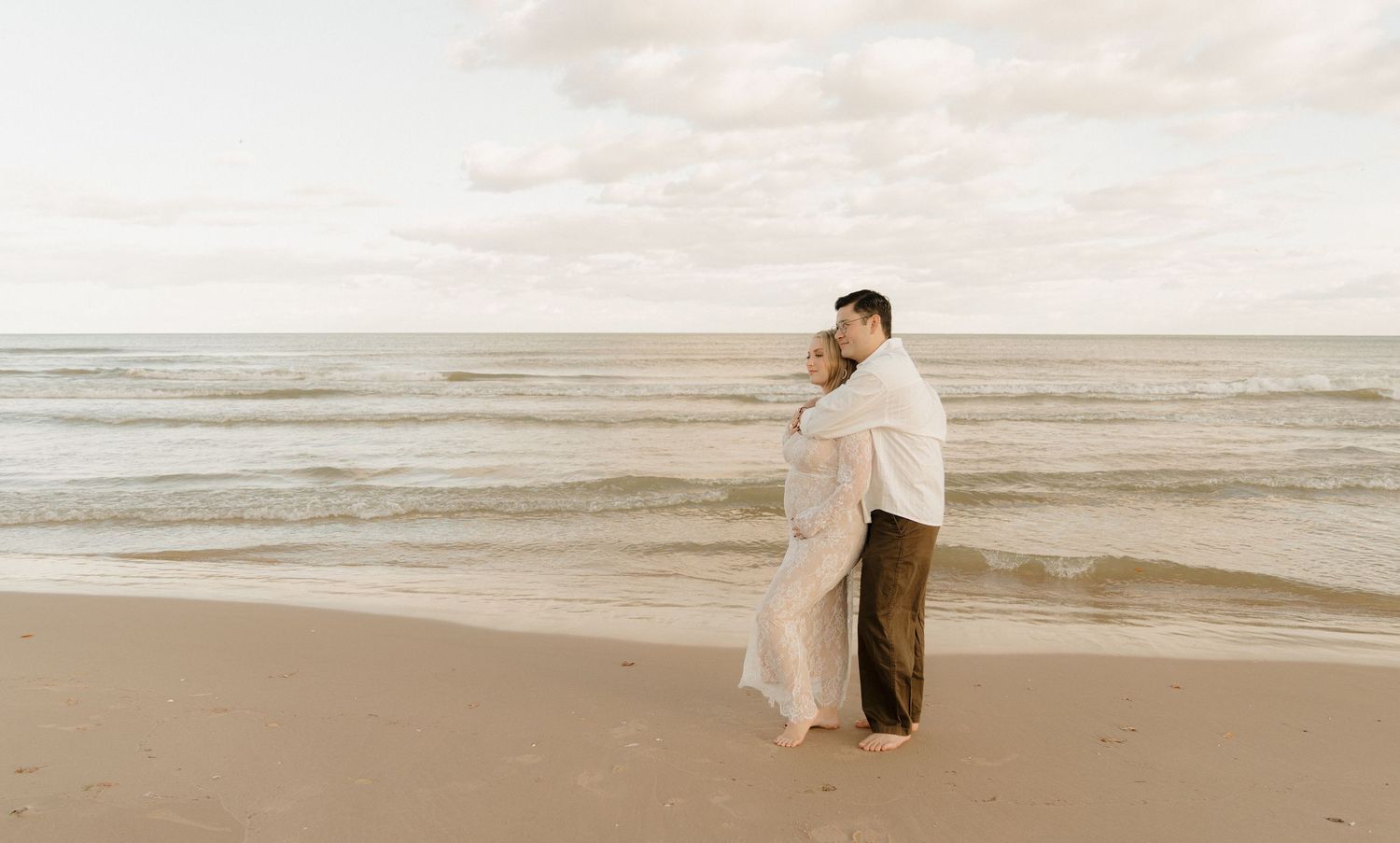 Couple embracing on a sandy beach with calm ocean waves and a soft cloudy sky in the background.