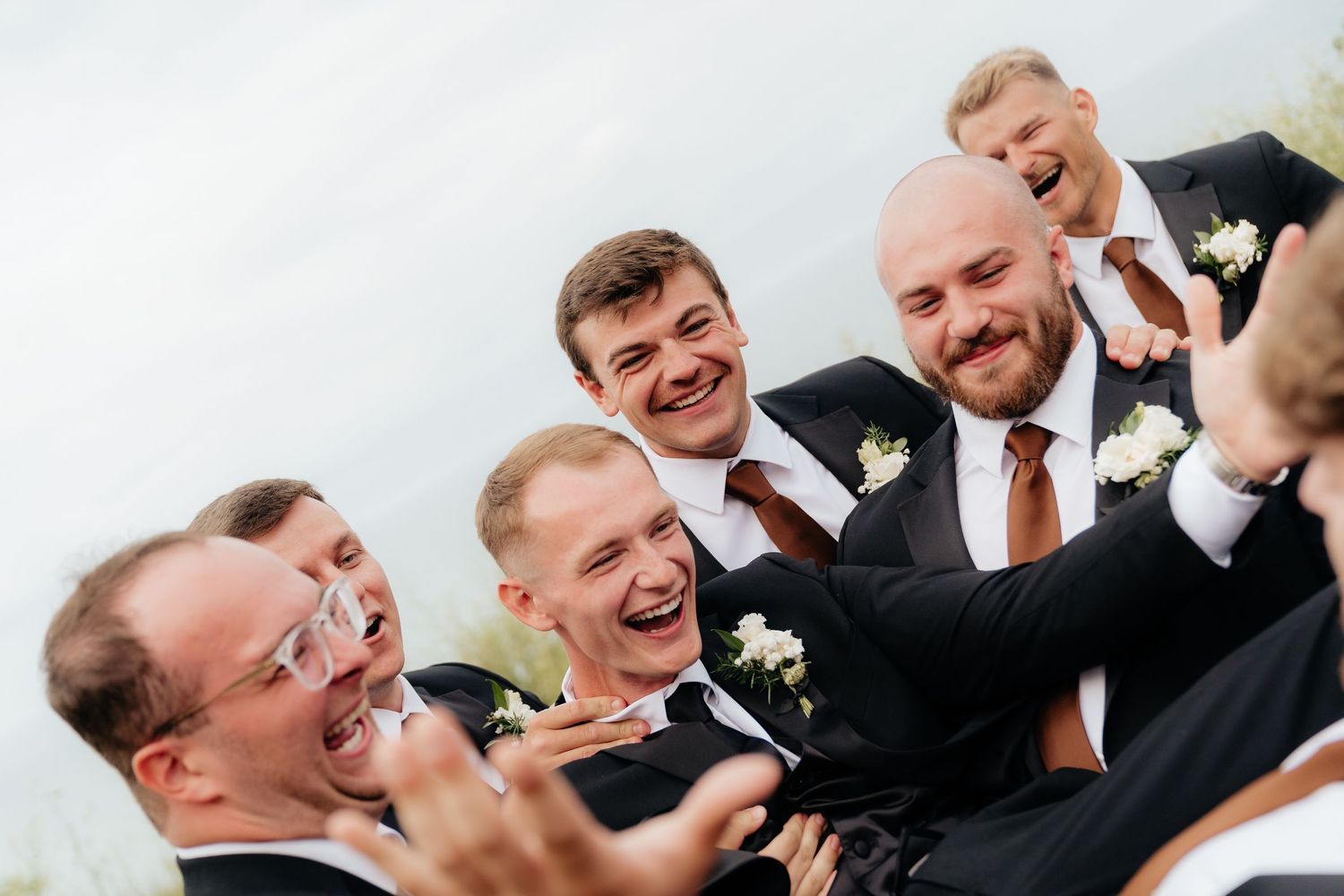 Groomsmen in dark suits laughing and celebrating together at a wedding, wearing white boutonnieres.