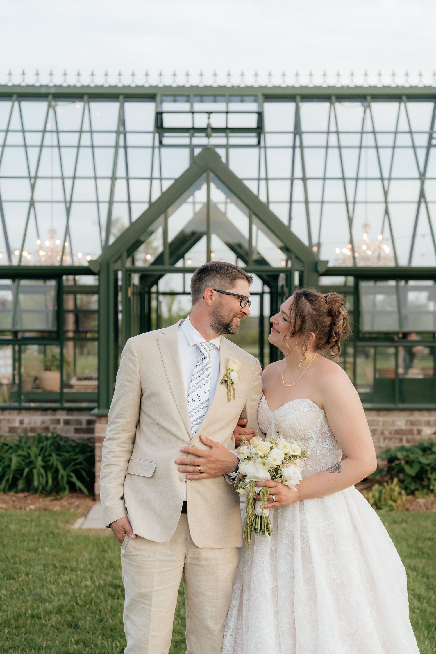 Bride and groom smiling at each other in front of a glass greenhouse, bride holding white bouquet, groom in tan suit.