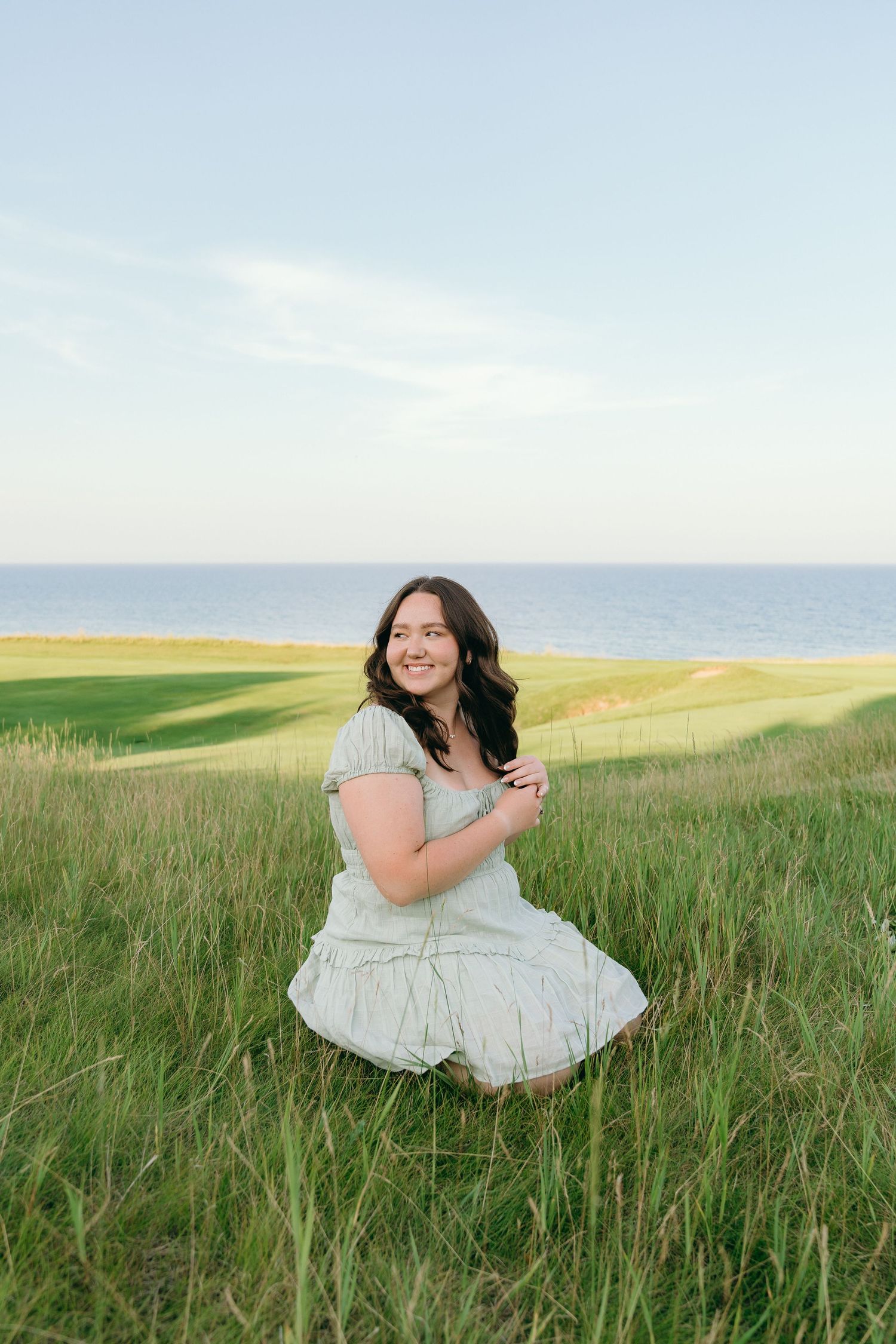 A smiling young woman sitting in a grassy field with the ocean in the background.