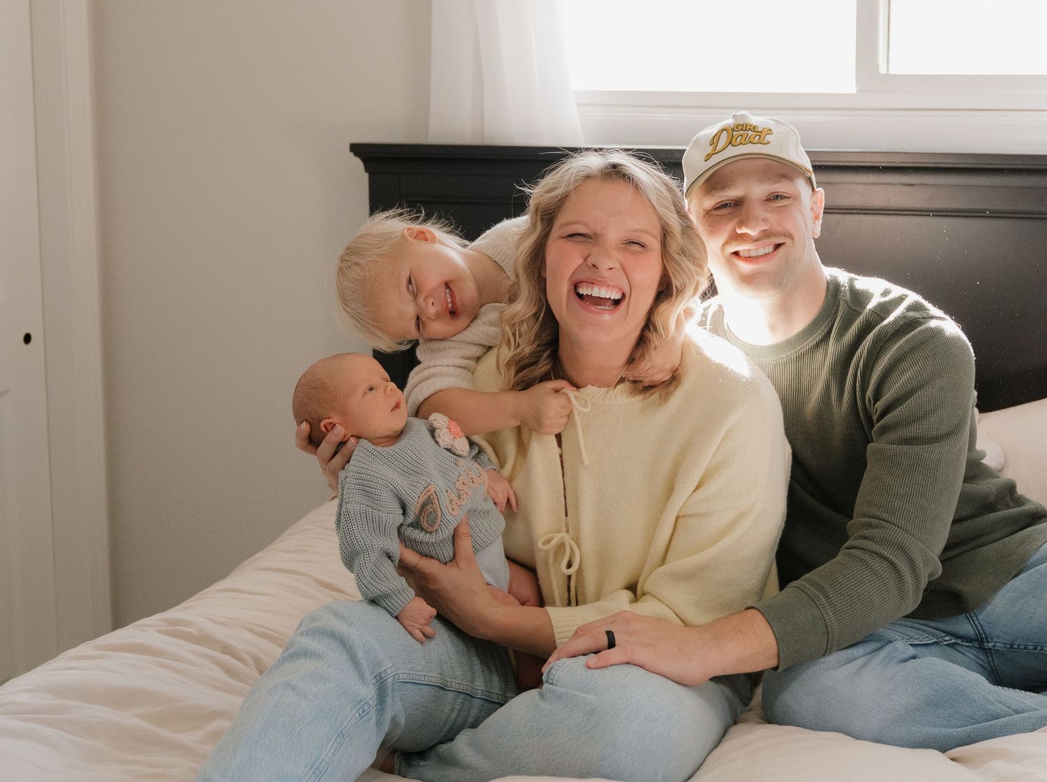 A smiling woman holding a baby while a young boy sits next to her on a bed.