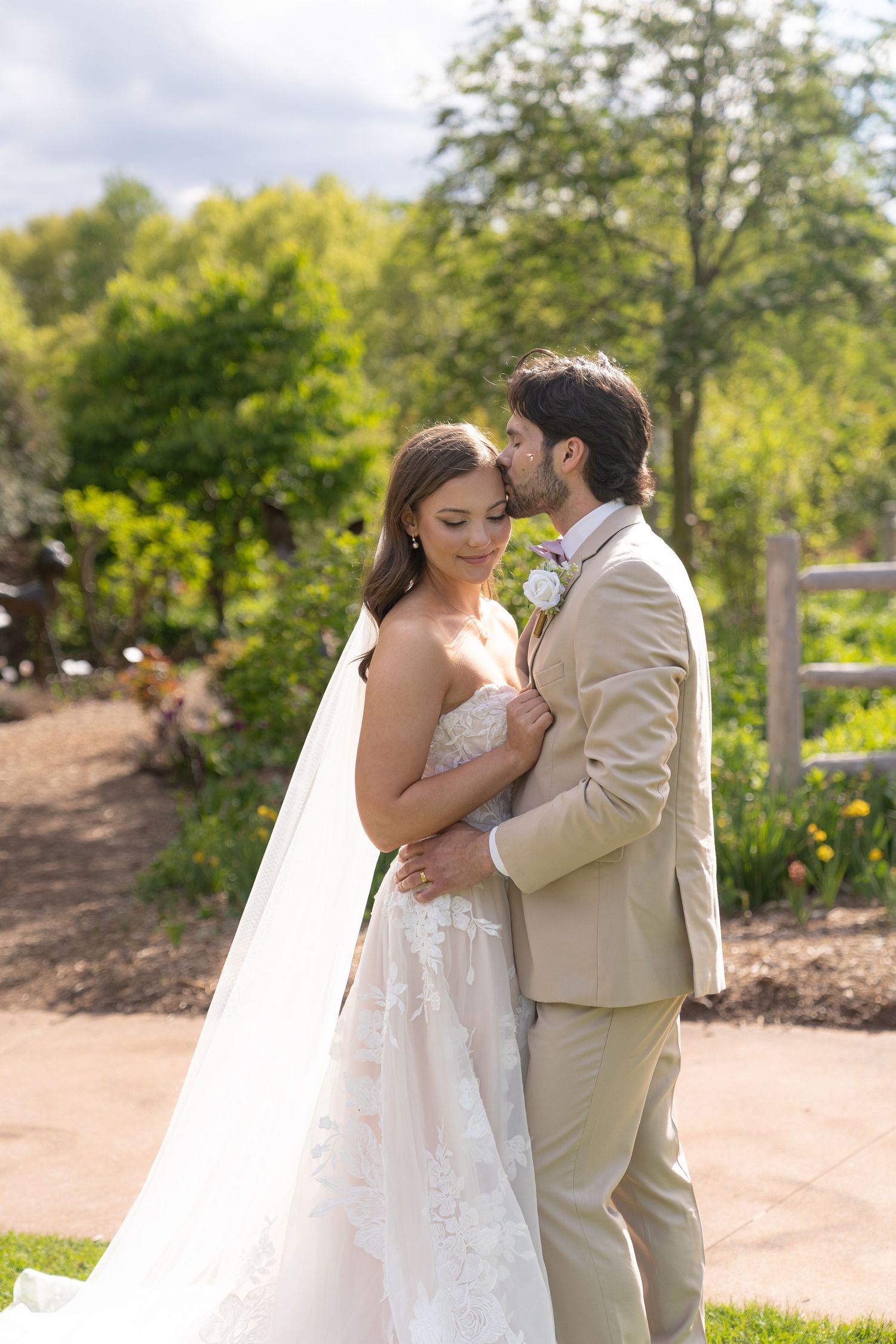 A bride and groom embrace in a garden setting with trees and foliage in the background.