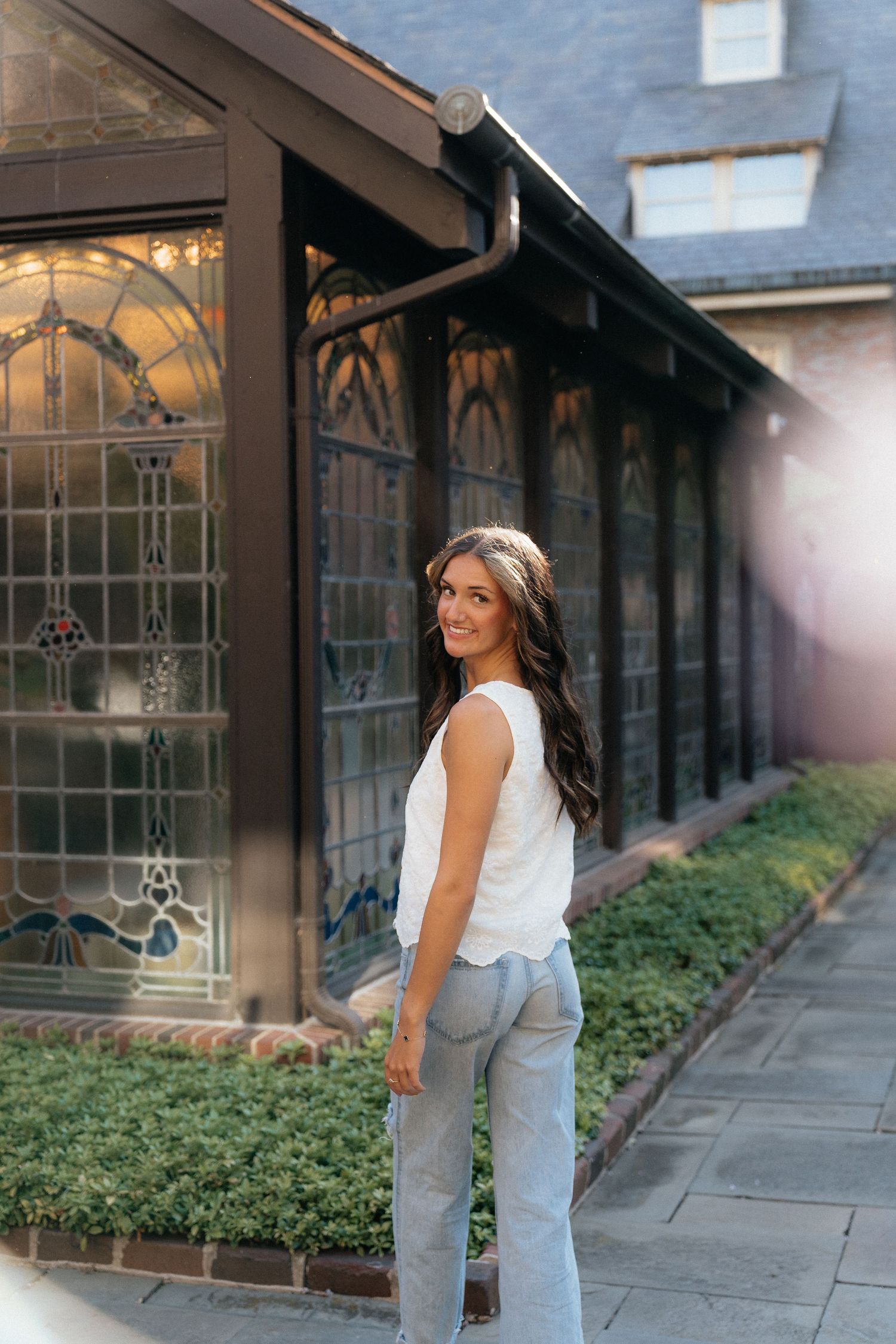 A smiling woman in white top and gray jeans poses outdoors beside stained glass windows of an elegant building.