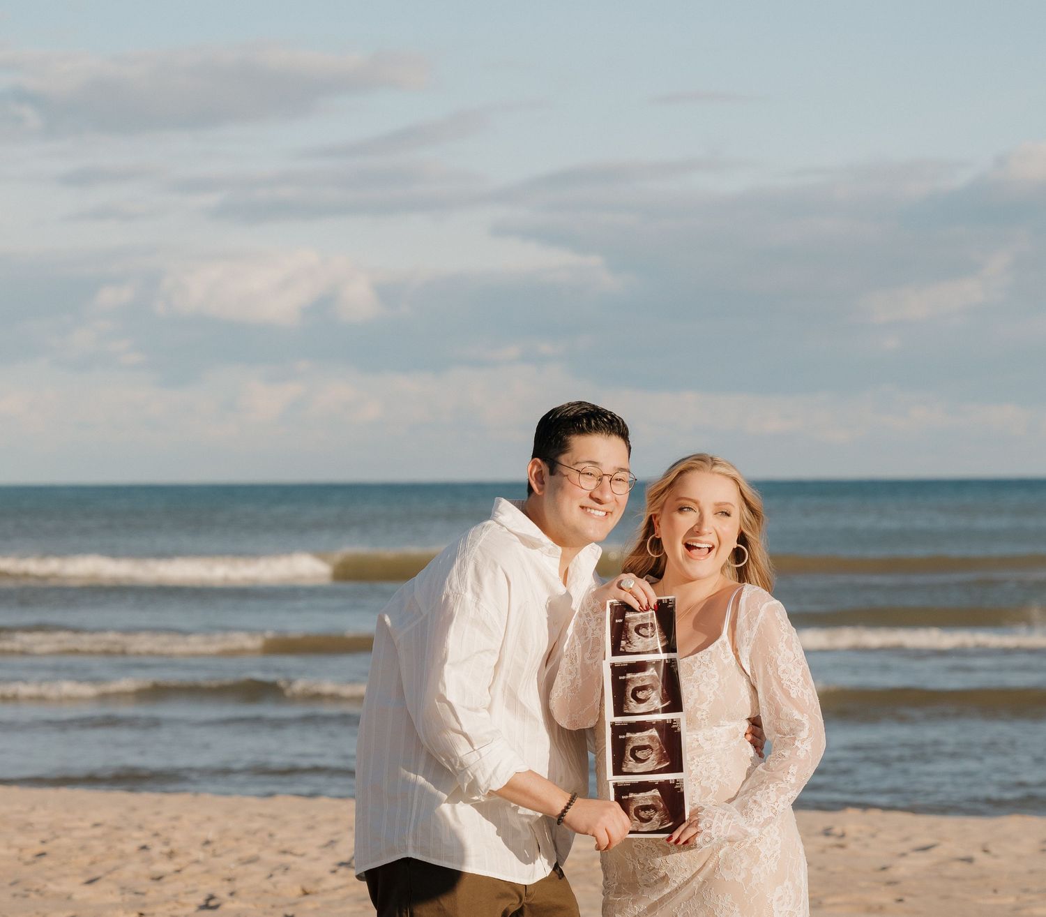Happy couple on beach holding ultrasound photos, pregnancy announcement, ocean backdrop, sunny day.