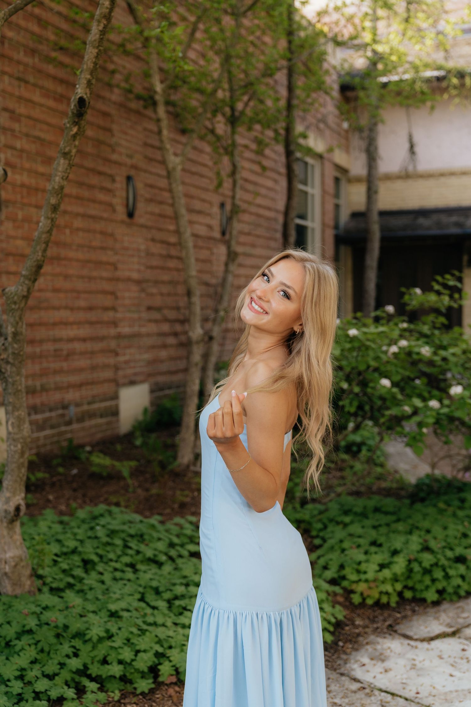 Smiling young woman in a light blue sundress posing outdoors near brick building with lush green garden.
