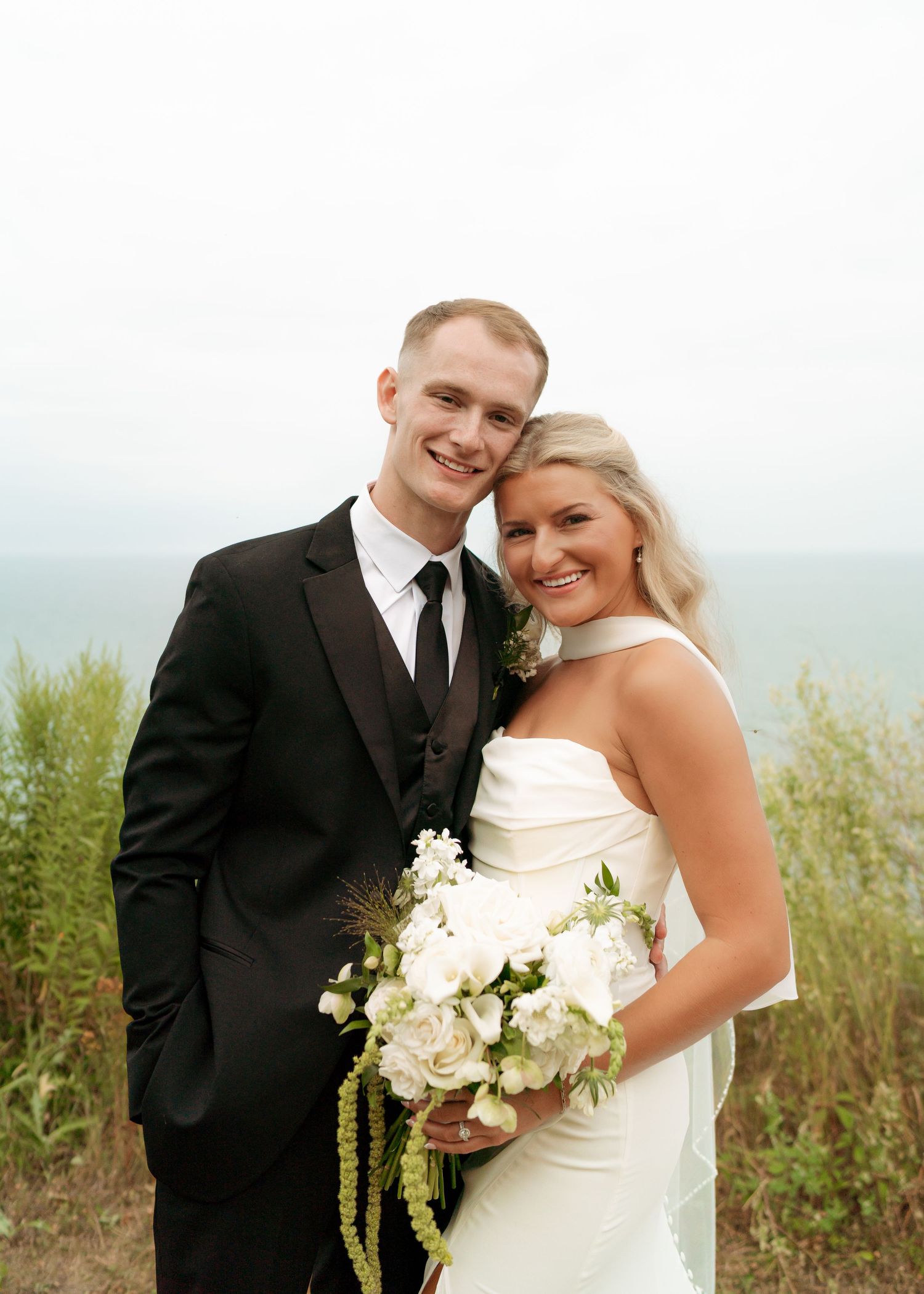Happy bride and groom pose outdoors on their wedding day, with the bride holding a white floral bouquet near the ocean.