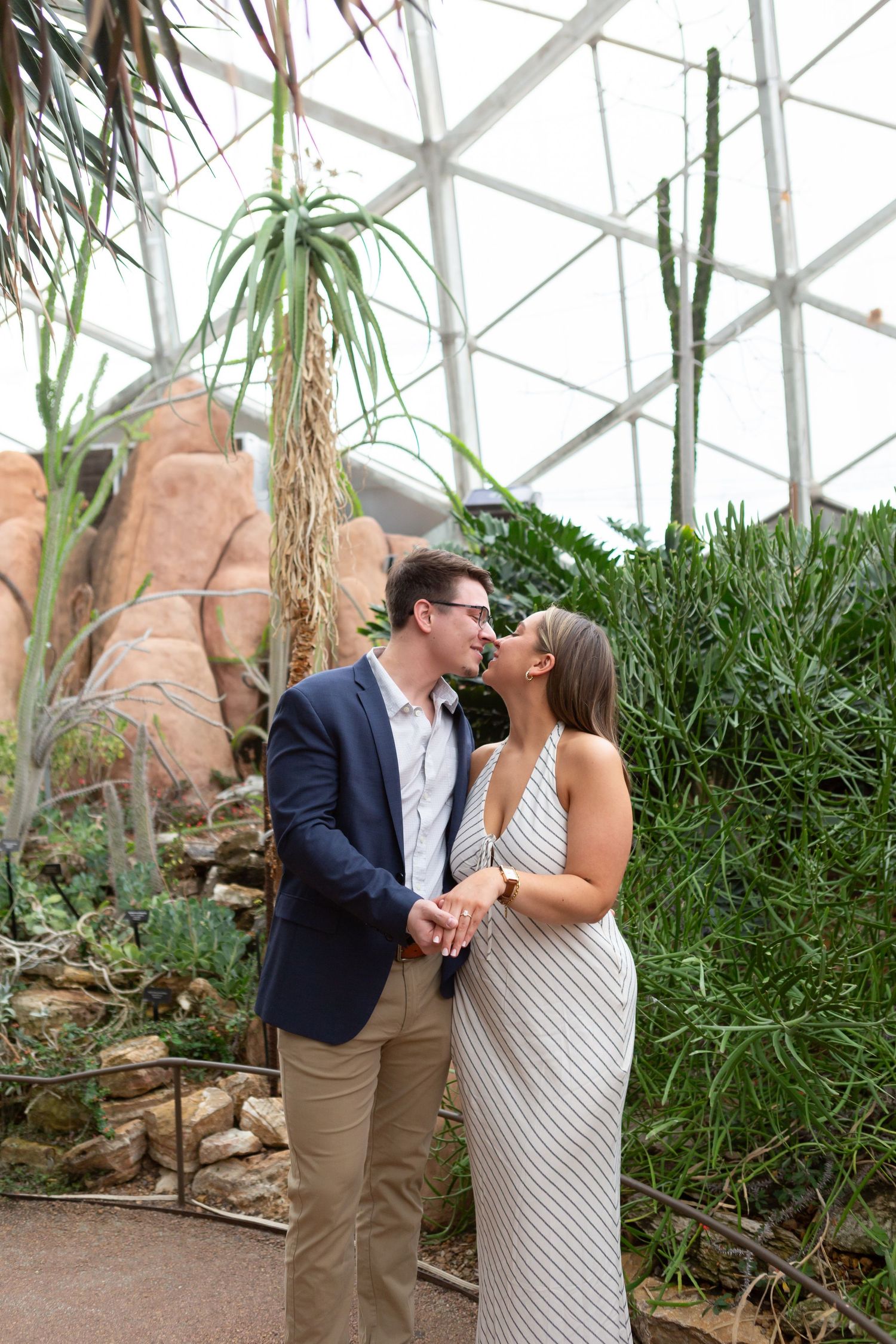 Couple holding hands and smiling at each other inside a botanical garden dome with cacti and succulents.