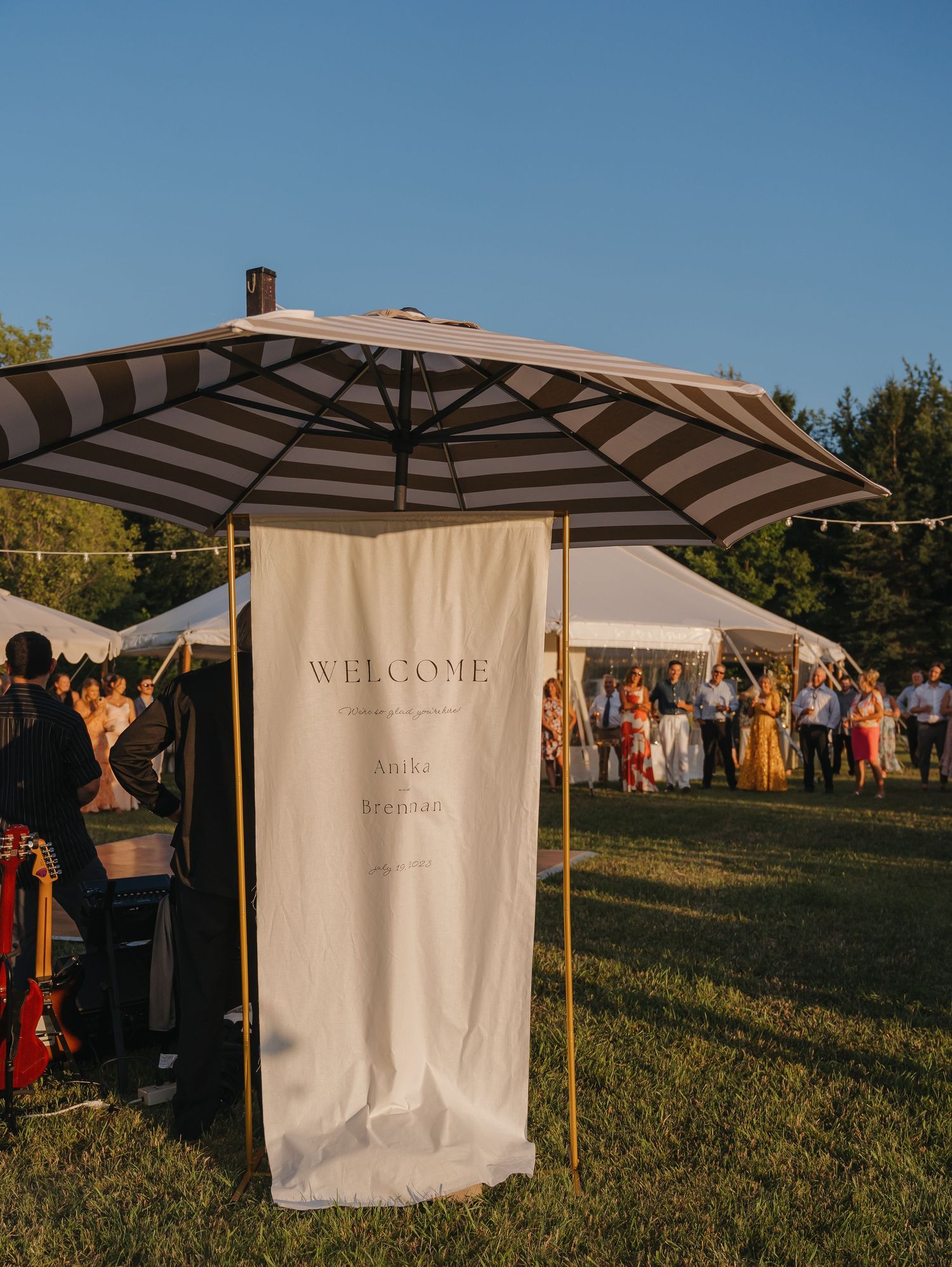 A large striped umbrella shades a welcome sign at an outdoor event or festival, with people gathered around.