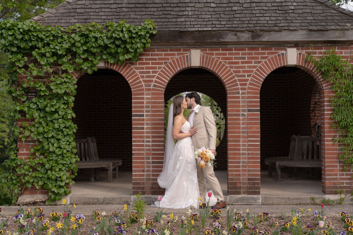A bride and groom stand together in a brick archway surrounded by lush greenery and flowers.
