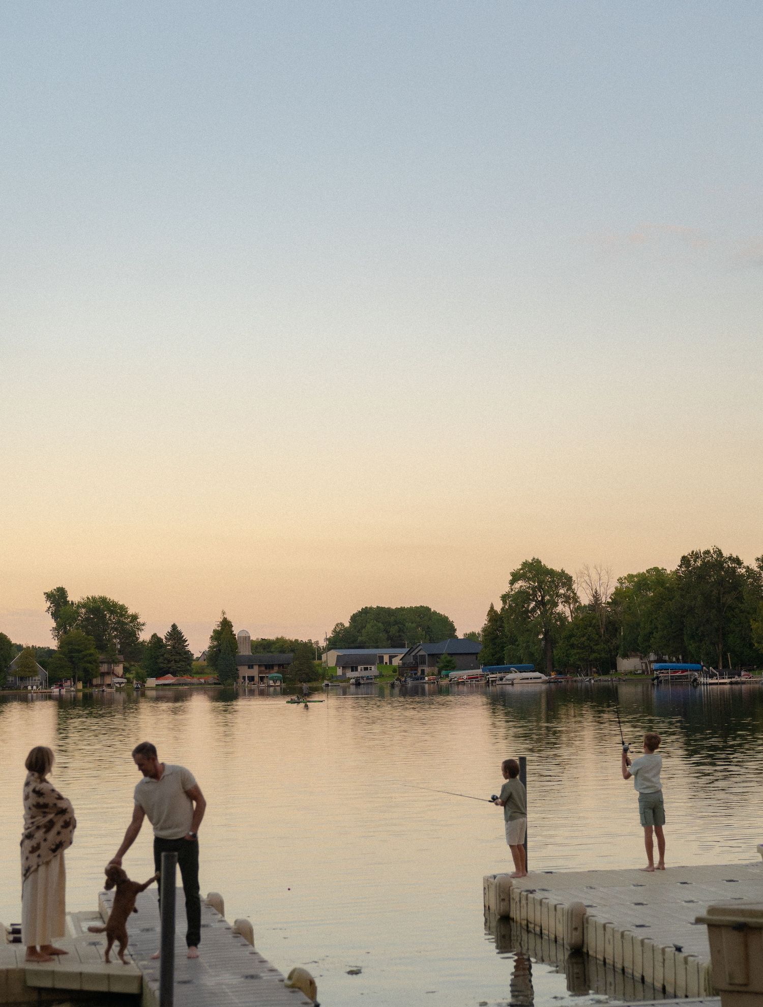 A scenic lakeside scene with people standing on a dock at sunset, surrounded by trees and buildings.